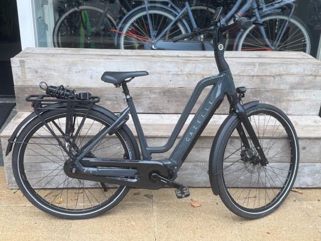 Donkergrijze Gazelle elektrische fiets geparkeerd voor een winkel, met spatborden, bagagedrager en schijfremmen.