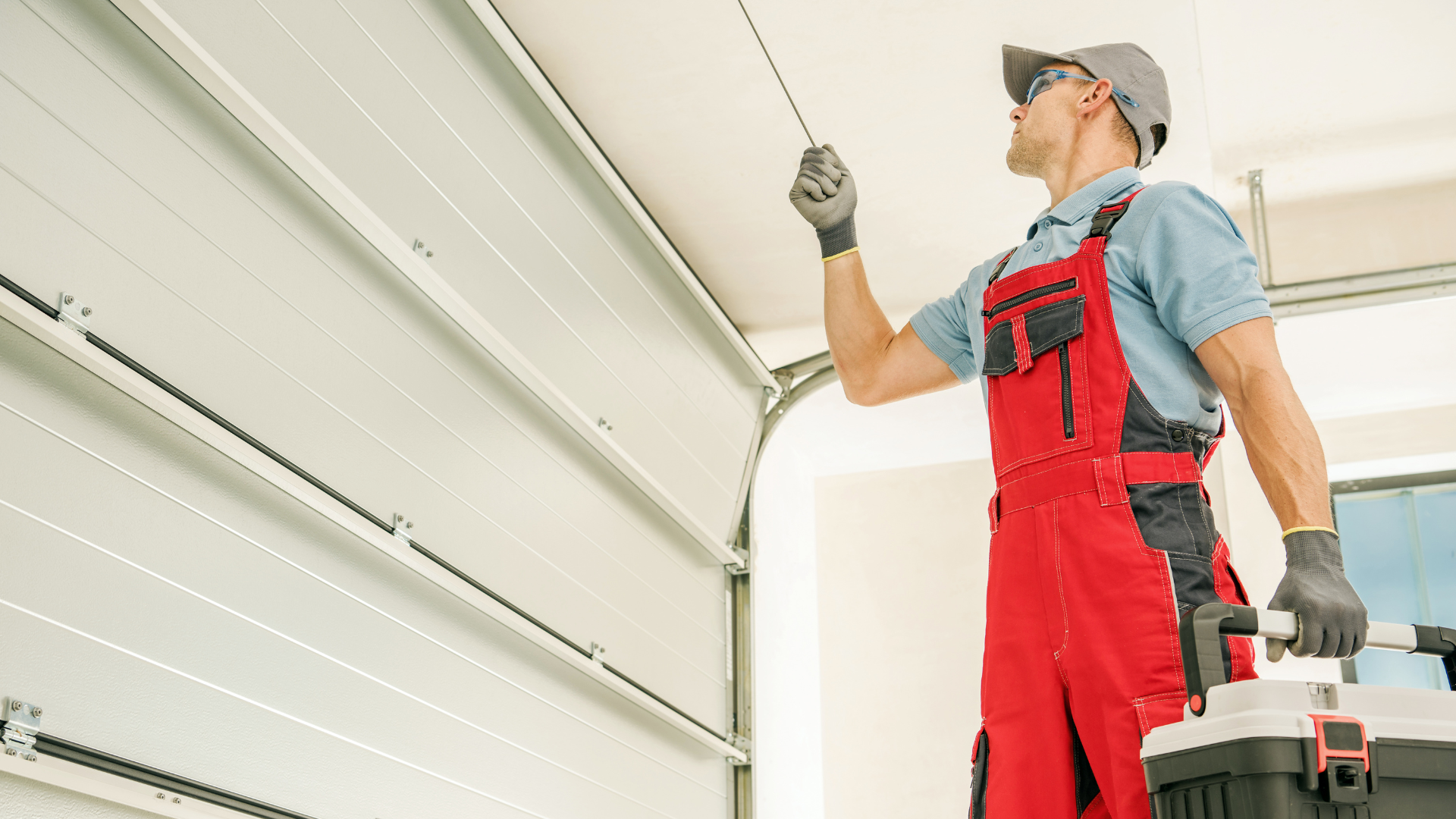Garage door repairman in red overalls and cap, inspecting overhead door with toolbox.