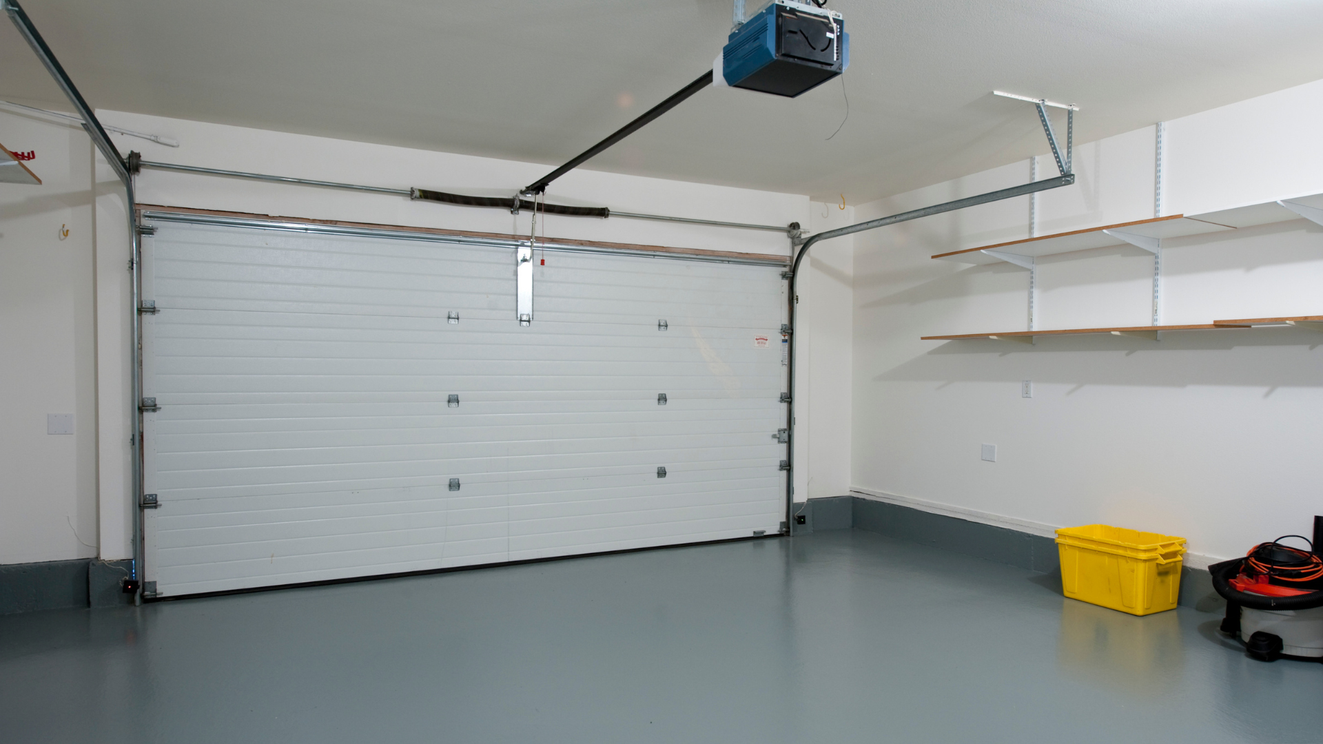 Inside a garage, a closed white door with gray floor, a yellow toolbox, and gray walls.