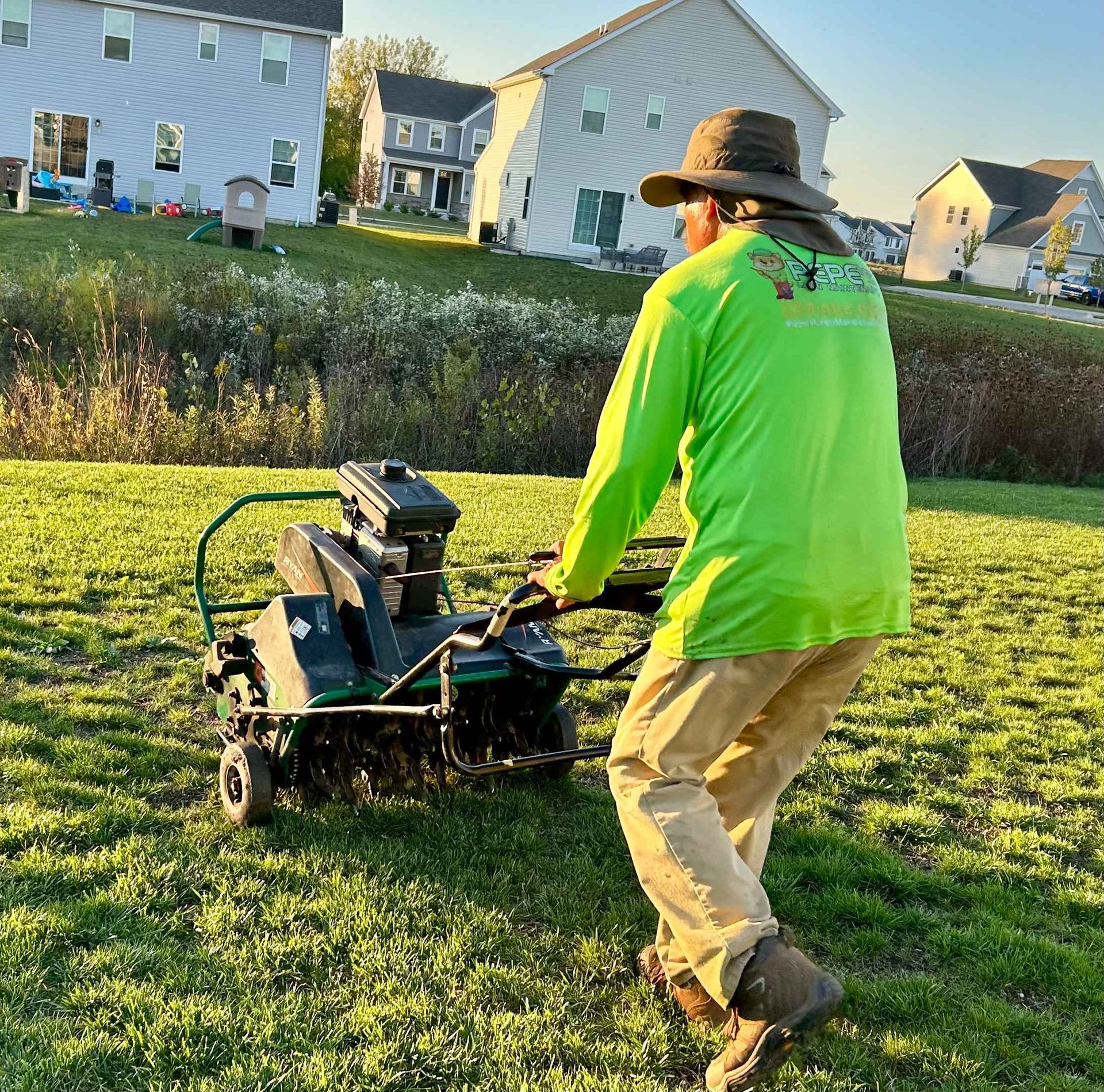 Man aerating a lawn with a machine. He's wearing a green shirt, tan pants, and a hat in a residential area.