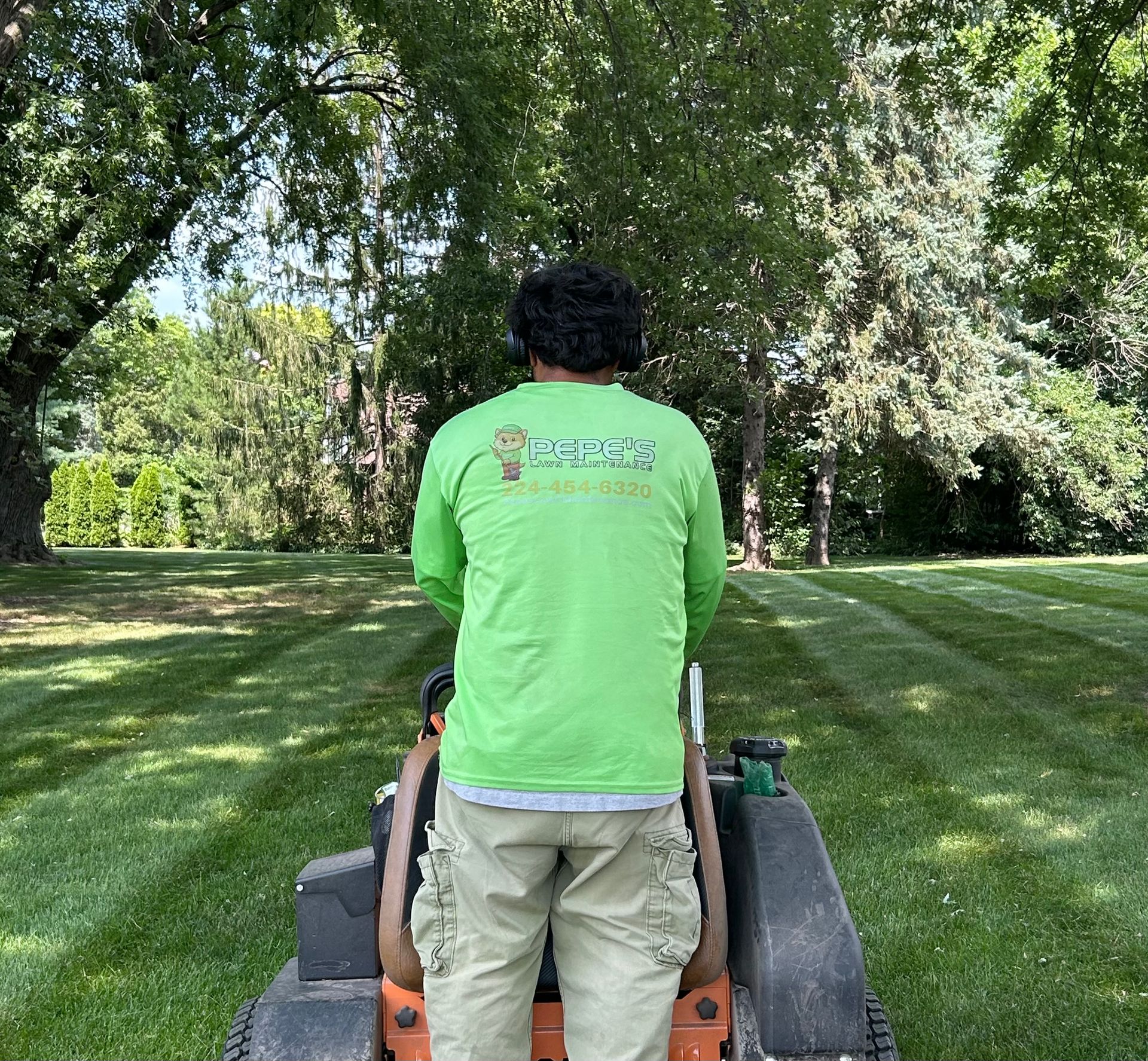 Person in a bright green shirt stands behind a mower on a grassy lawn with trees in the background