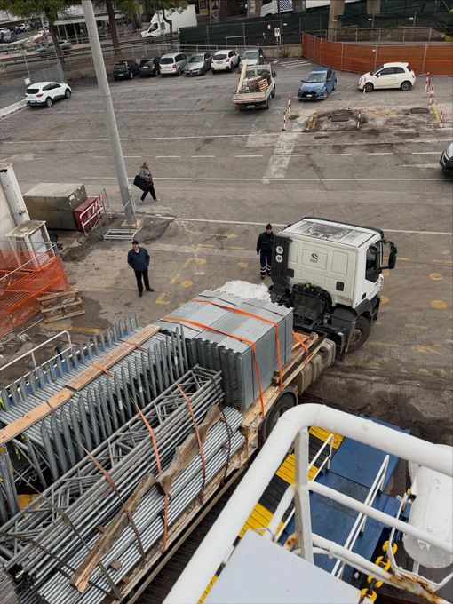 Un camion da cantiere e degli operai in un cantiere all'aperto e bagnato, accanto a travi metalliche impilate e auto parcheggiate.