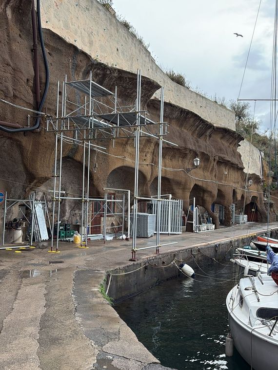 Cantiere edile a picco sul mare, accanto a un porto, con impalcature e barche ormeggiate sottostanti.