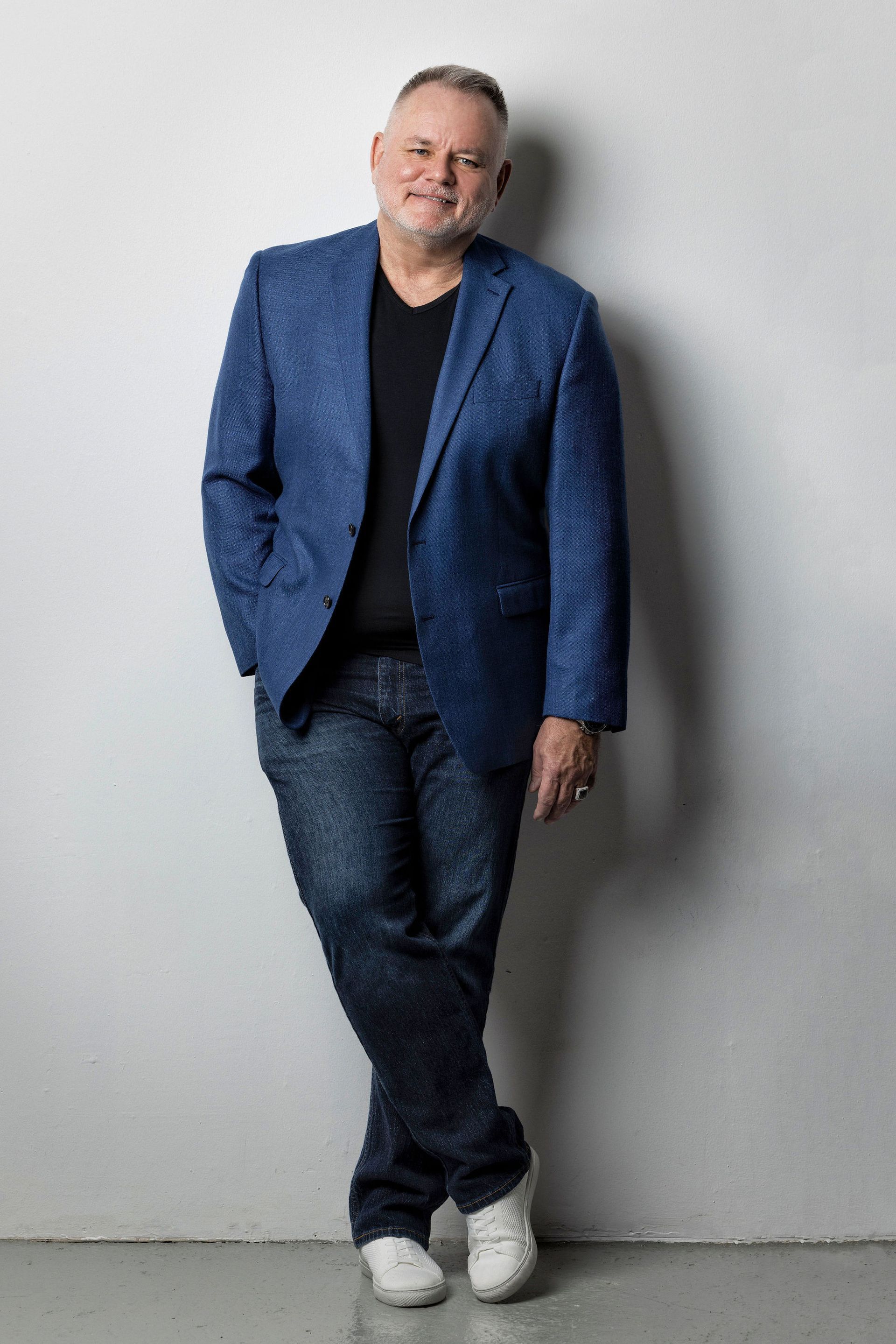 Man in a blue blazer, black shirt and jeans, leaning against a white wall with white sneakers.