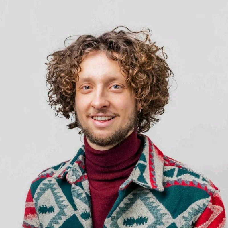 Portretfoto van een glimlachende man met krullend bruin haar, een korte baard en een kleurrijke jas over een bordeauxrode coltrui tegen een lichte achtergrond.