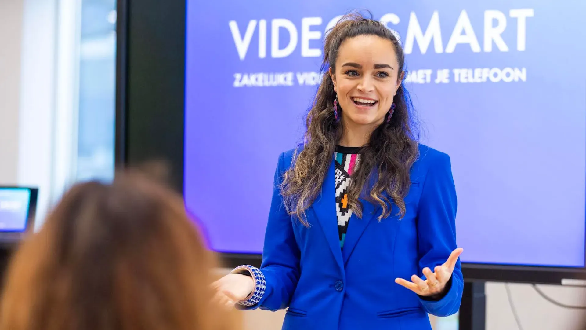 Een glimlachende persoon in een blauwe blazer staat voor een scherm met het opschrift VIDEOSMART en gebaart terwijl hij of zij een publiek toespreekt.