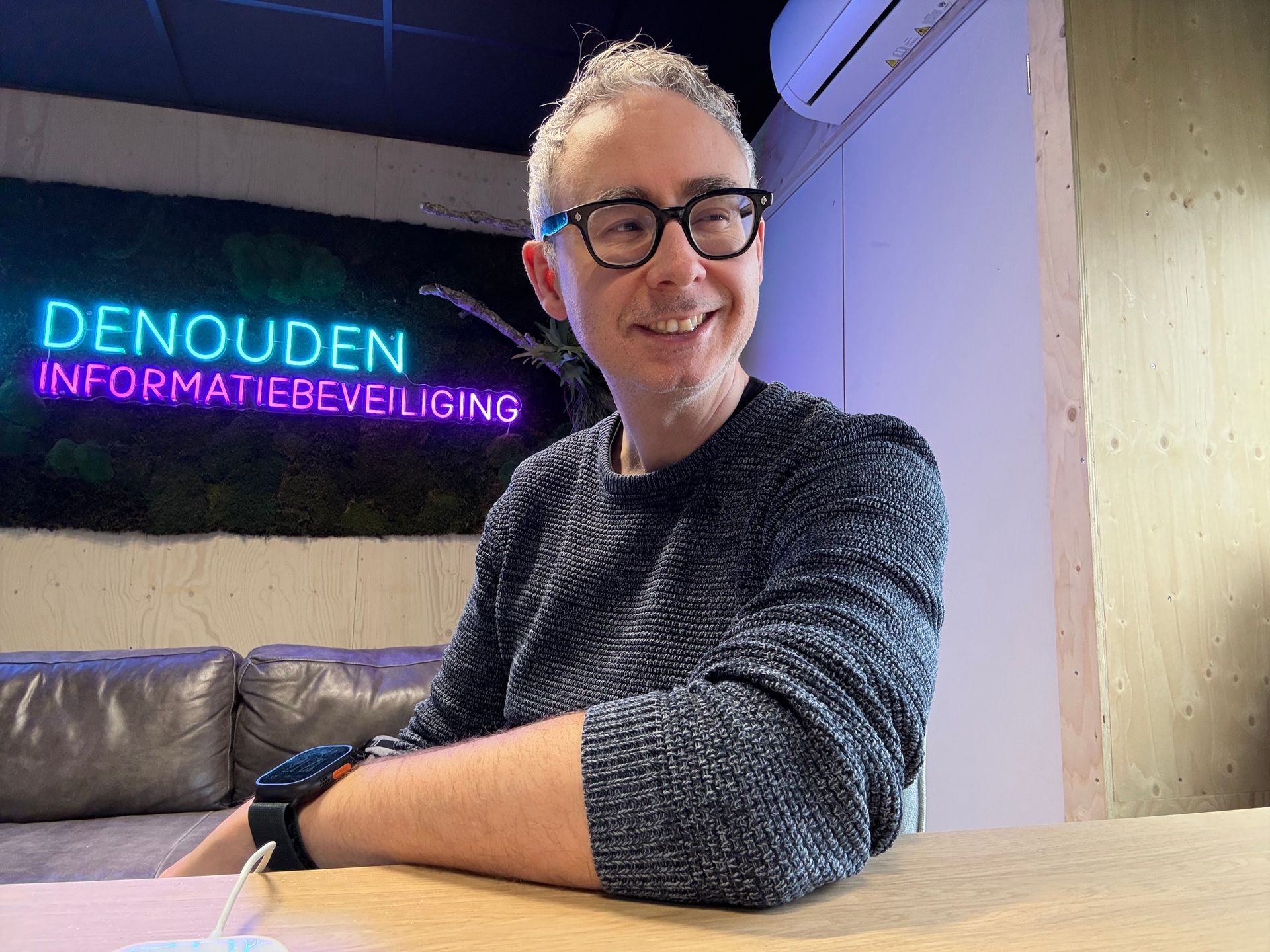 Een persoon in een grijze trui en met een bril zit aan een tafel vlakbij een muur met een neonreclame waarop staat