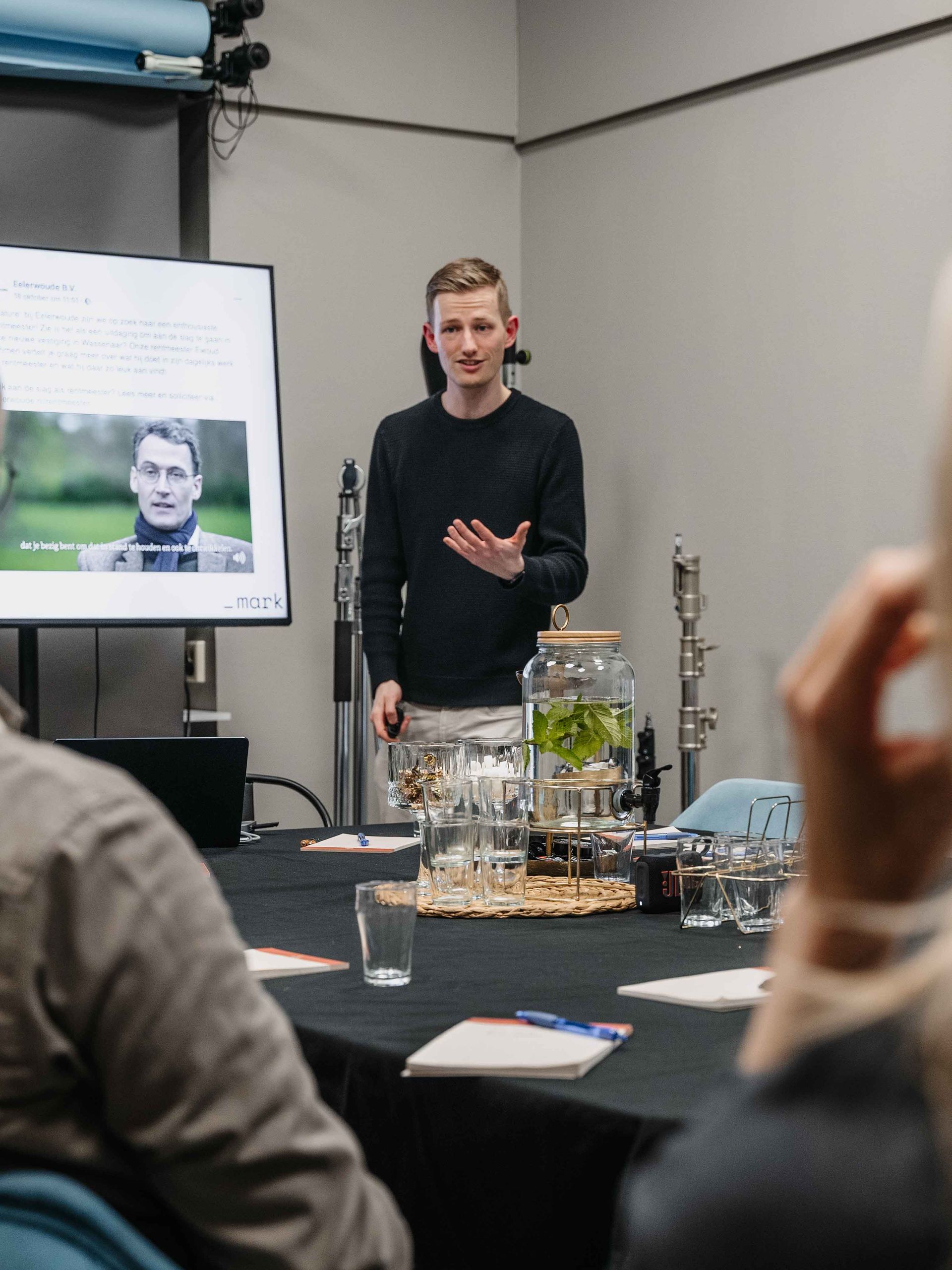 Tijdens een presentatie in een vergaderruimte maakt een spreker gebaren, staand naast een monitor waarop zijn of haar foto aan het publiek wordt getoond.