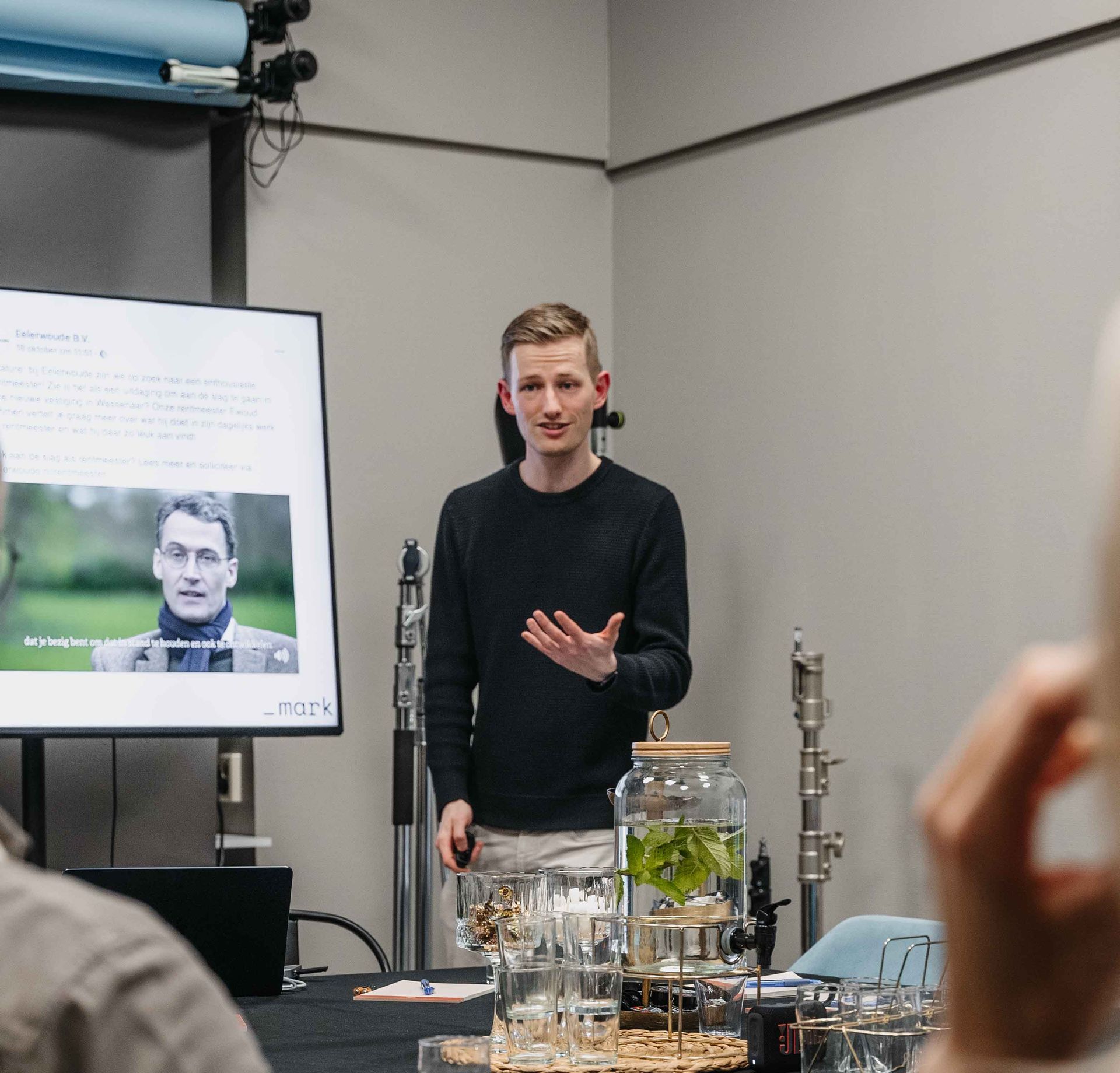 Een spreker gebaart terwijl hij een presentatie geeft voor een scherm waarop een foto van hemzelf in een studio-omgeving te zien is.