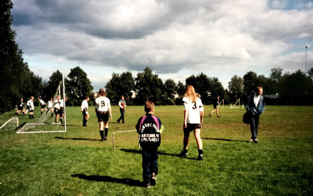 Een groep mensen in sportshirts loopt over een grasveld bij een voetbaldoel onder een bewolkte hemel.