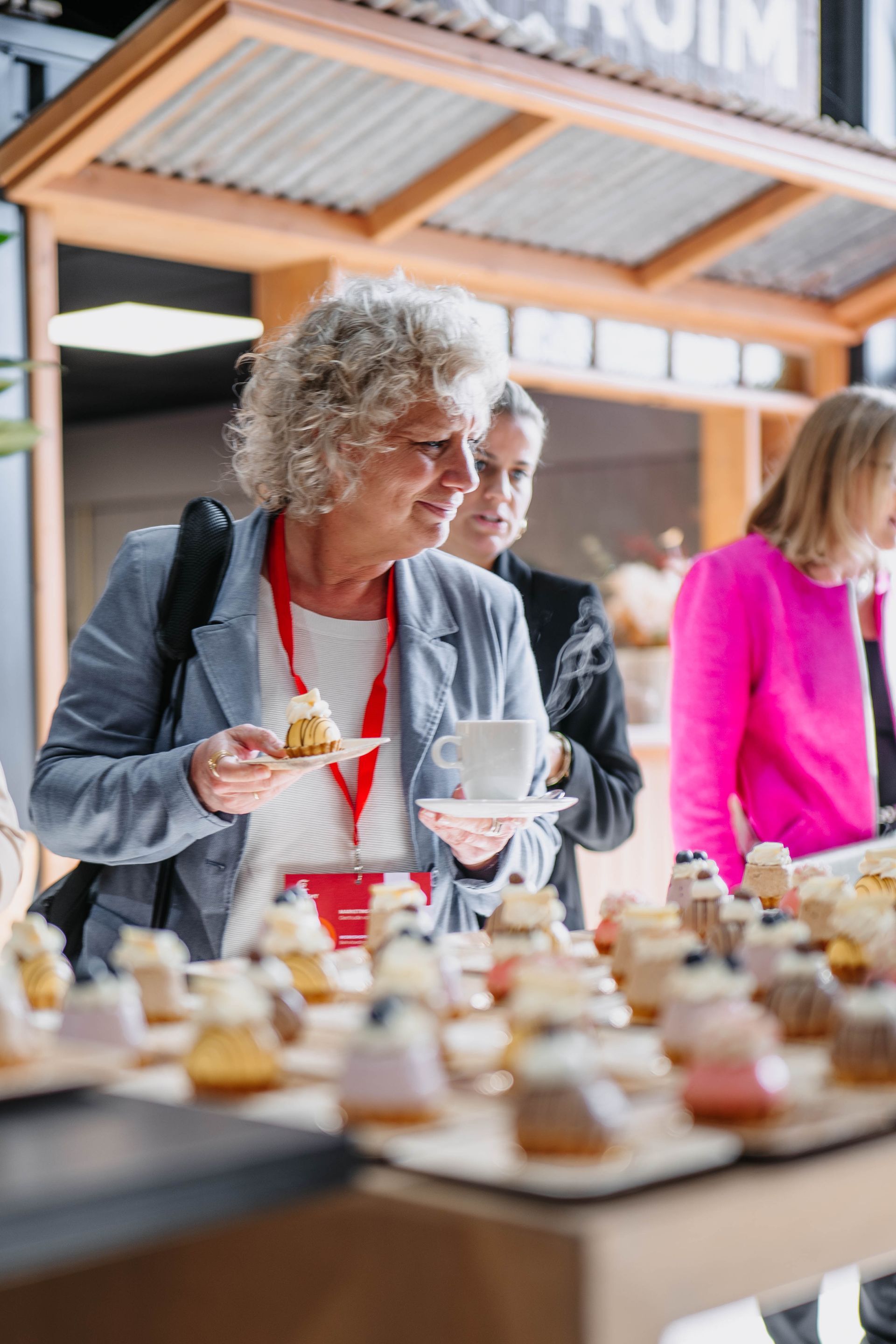 Een persoon glimlacht terwijl hij een klein dessert en een kopje vasthoudt aan een tafel vol hapjes in een café.