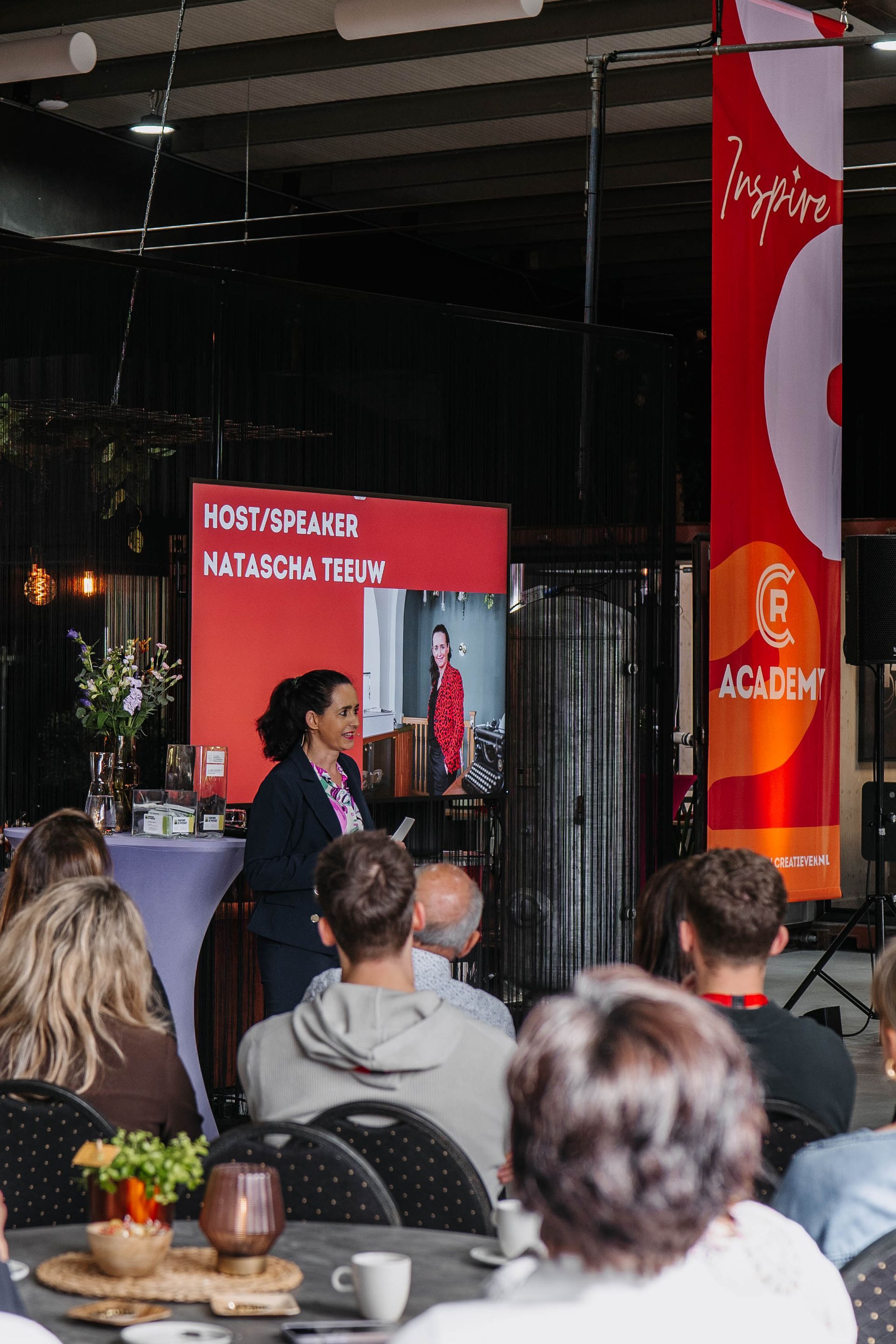 Een spreker spreekt een publiek toe in een zaal met een groot rood beeldscherm en een verticaal oranje en rood spandoek.