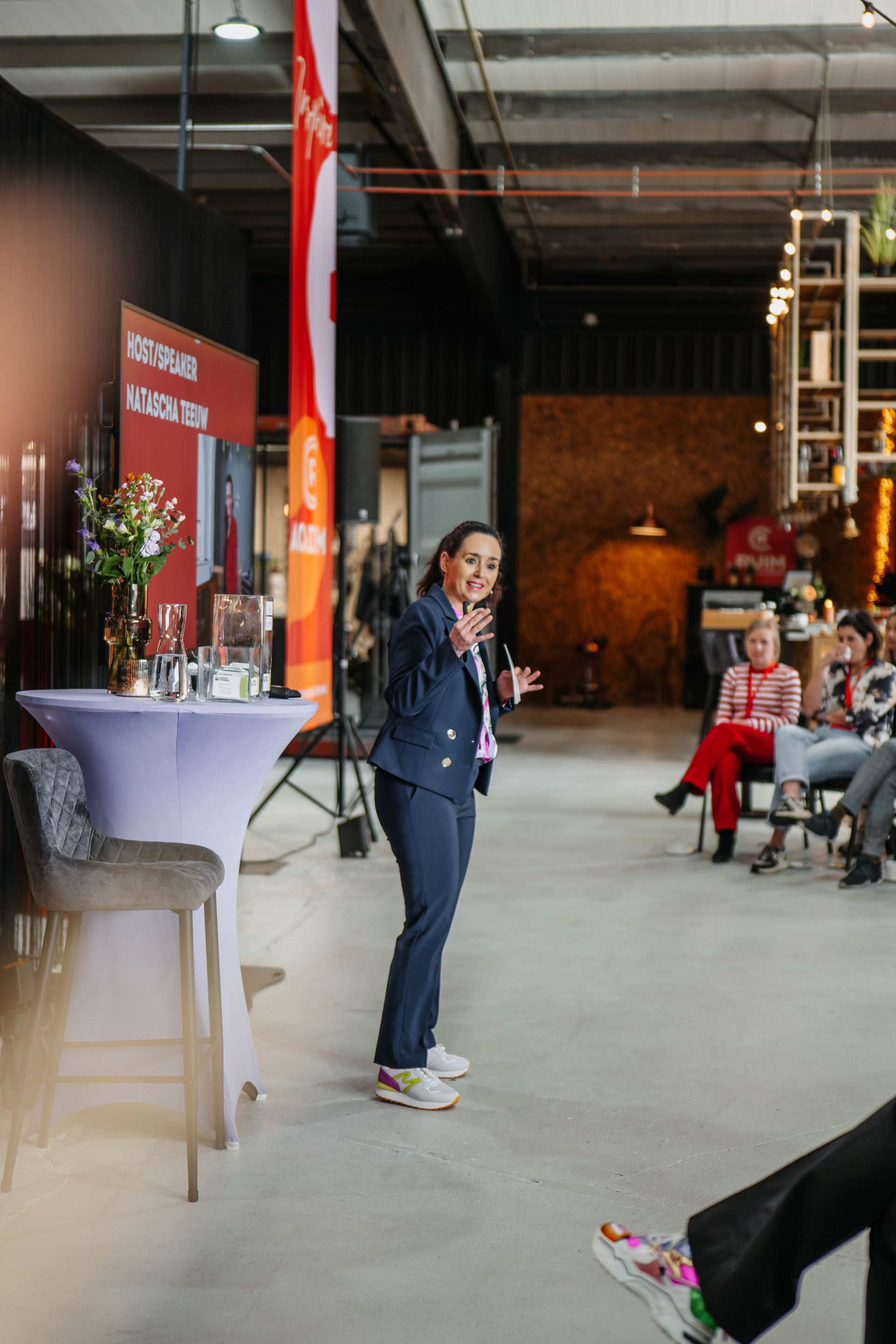 Een persoon in een blauw pak gebaart terwijl hij een publiek toespreekt in een ruime, industriële zaal met evenementborden.