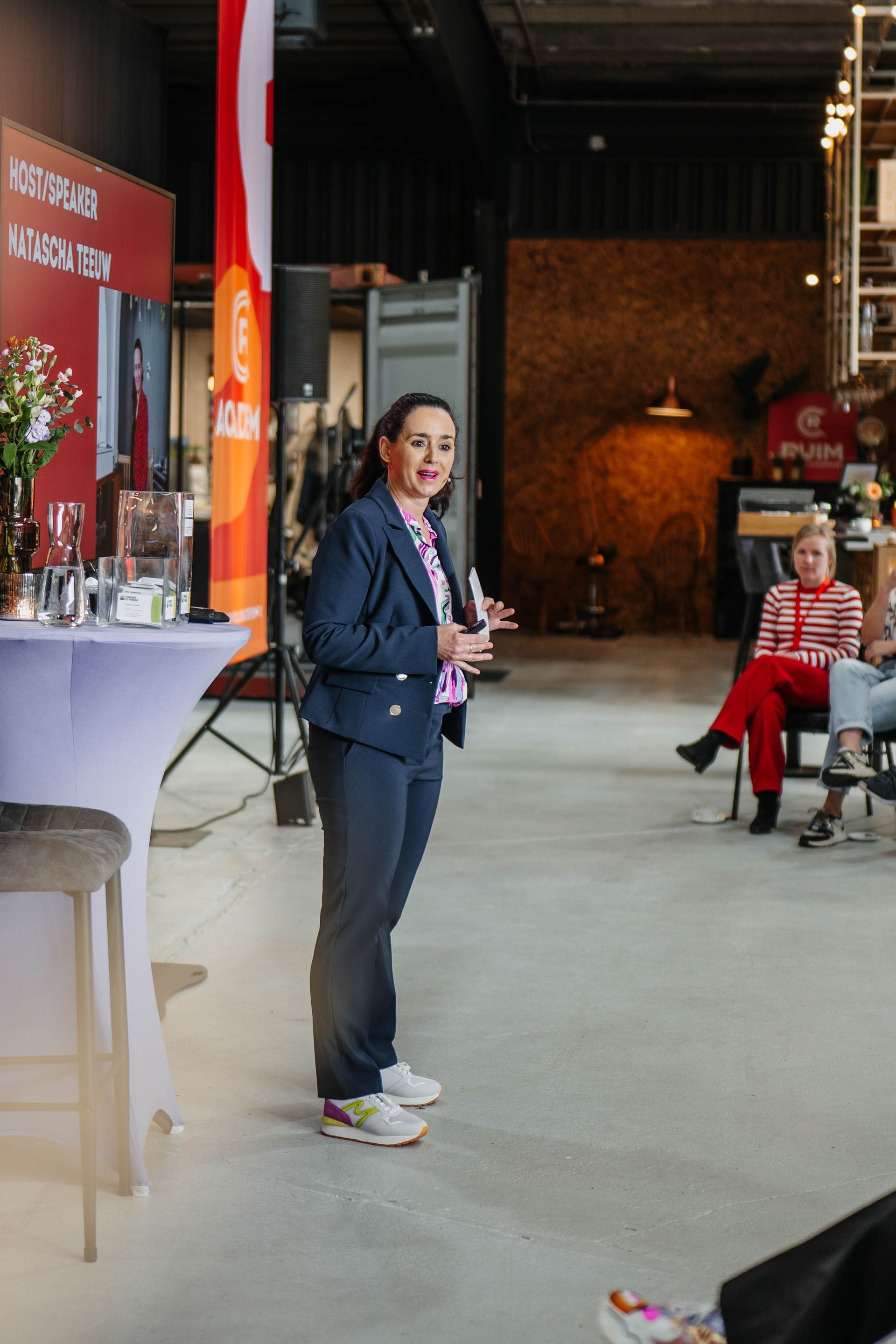 Een persoon in een donkere blazer en broek gebaart terwijl hij een publiek toespreekt in een ruime, industrieel ogende zaal.