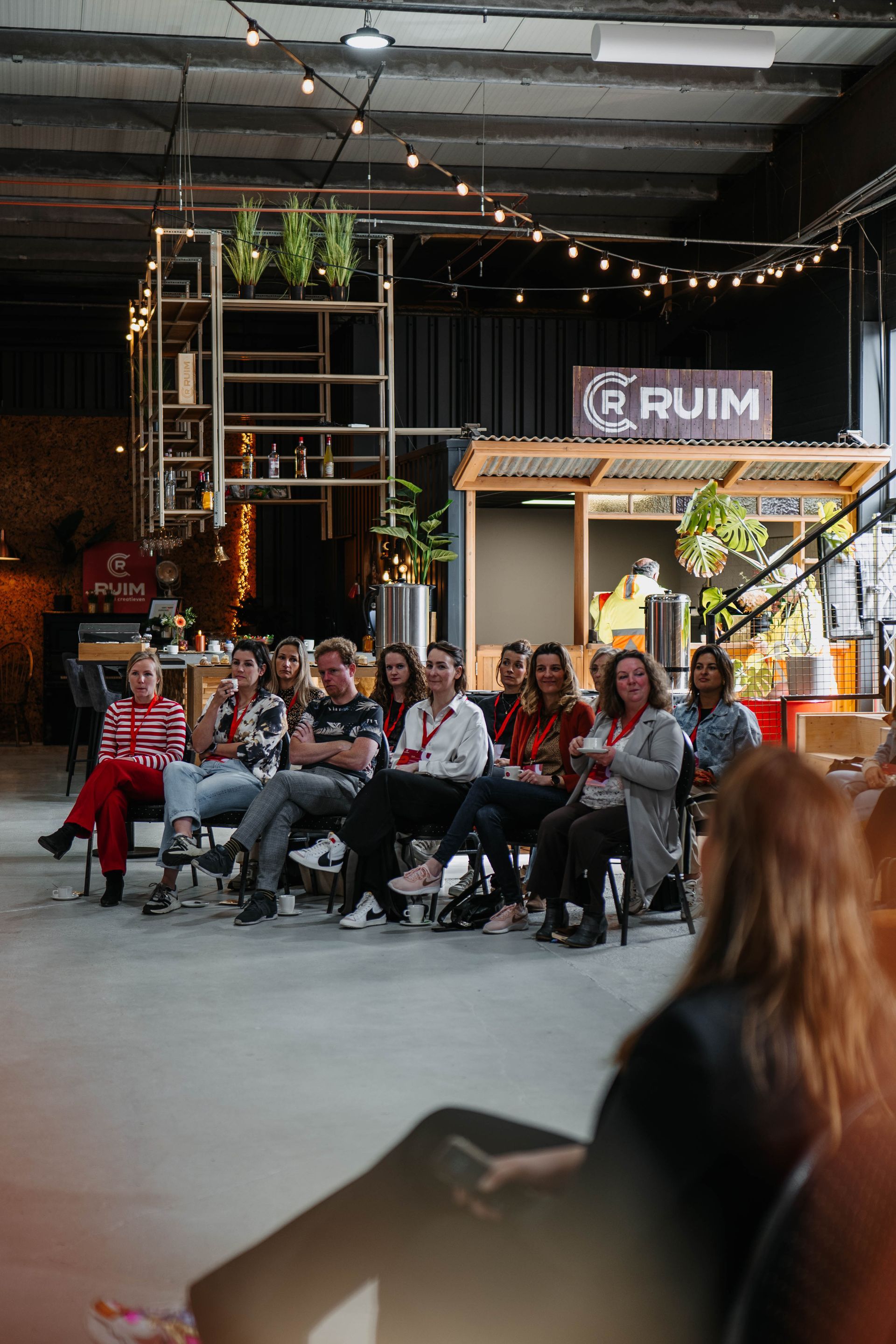 Een groep mensen zit in een ruimte in industriële stijl en luistert naar een presentatie in een overdekte evenementenruimte.