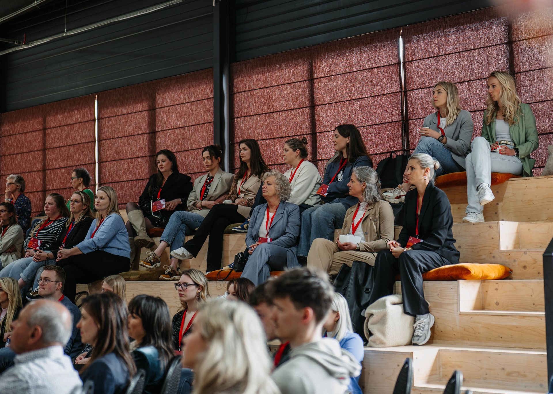 Een groep mensen zit op houten tribunebanken in een grote zaal en kijkt naar een presentatie.