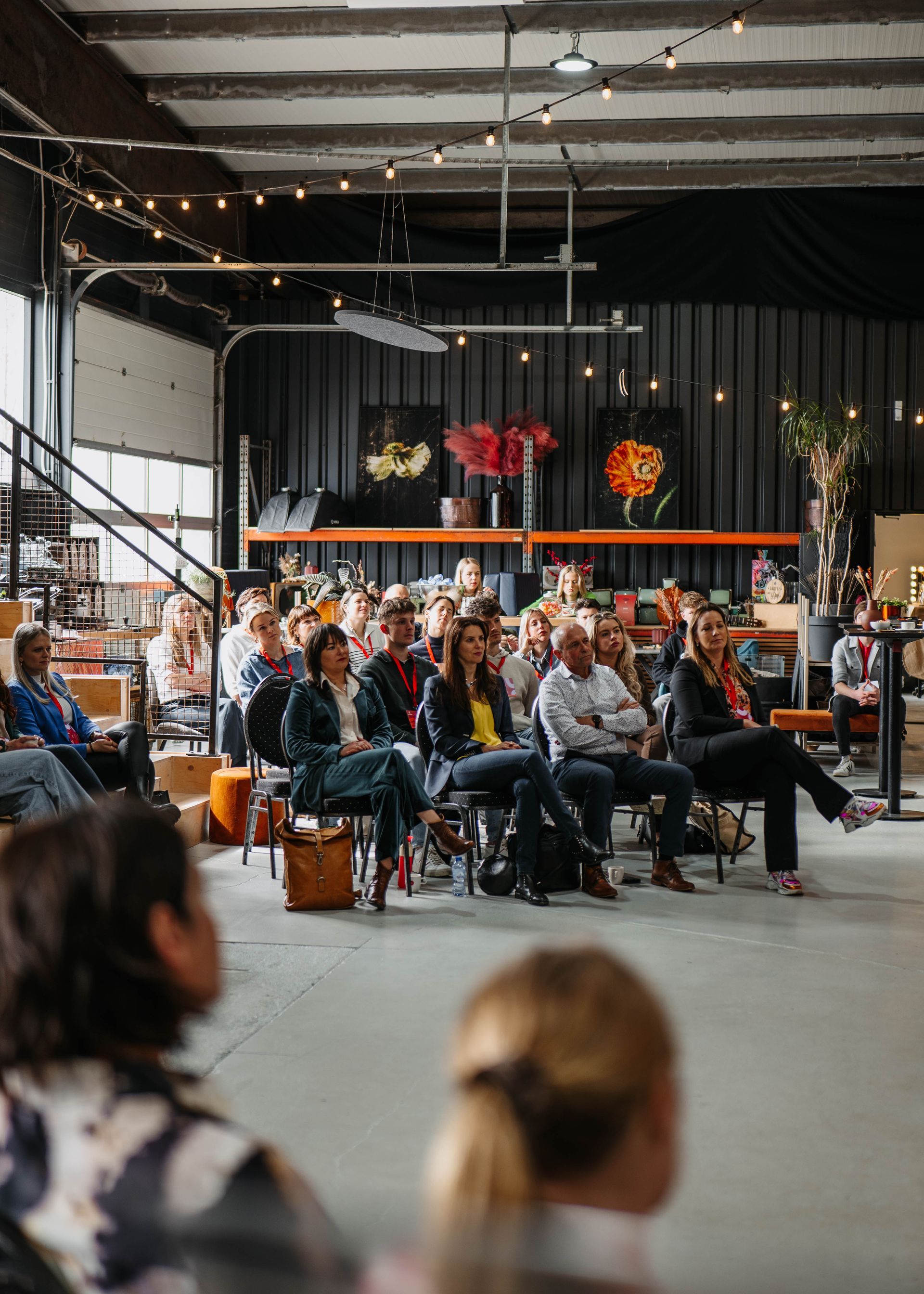 Een groep mensen zit in een evenementenruimte in industriële stijl, met hun gezicht naar voren gericht, terwijl ze naar een presentatie luisteren.