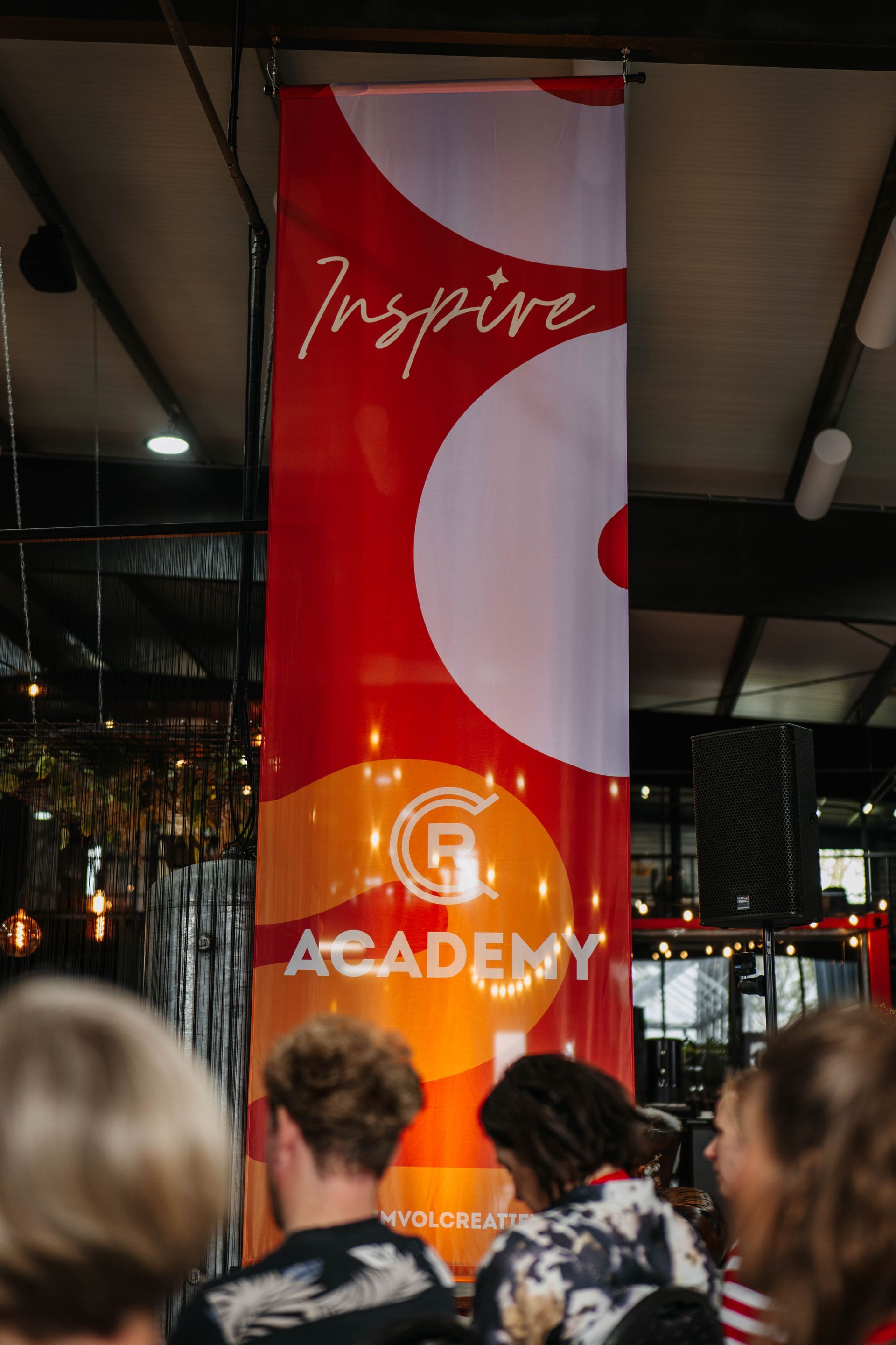 In een zaal achter een publiek hangt een verticaal rood en oranje spandoek met de tekst 