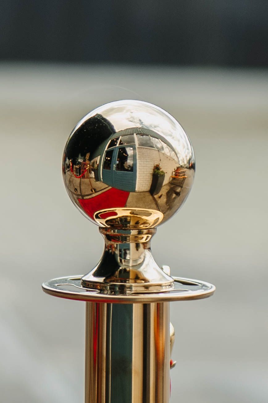 Een close-up van een gepolijste gouden afzetpaal met een bolvormige bovenkant, die een rood tapijt en de omringende omgeving weerspiegelt.