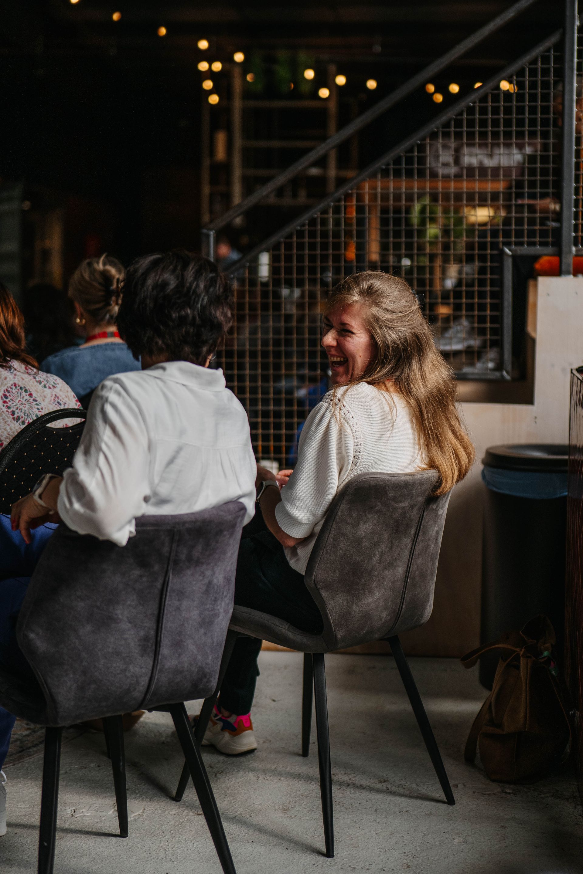 Twee mensen zitten op stoelen in een schemerige, met lichtslingers verlichte ruimte; de ​​persoon aan de rechterkant is naar de persoon naast zich toegekeerd om te glimlachen.
