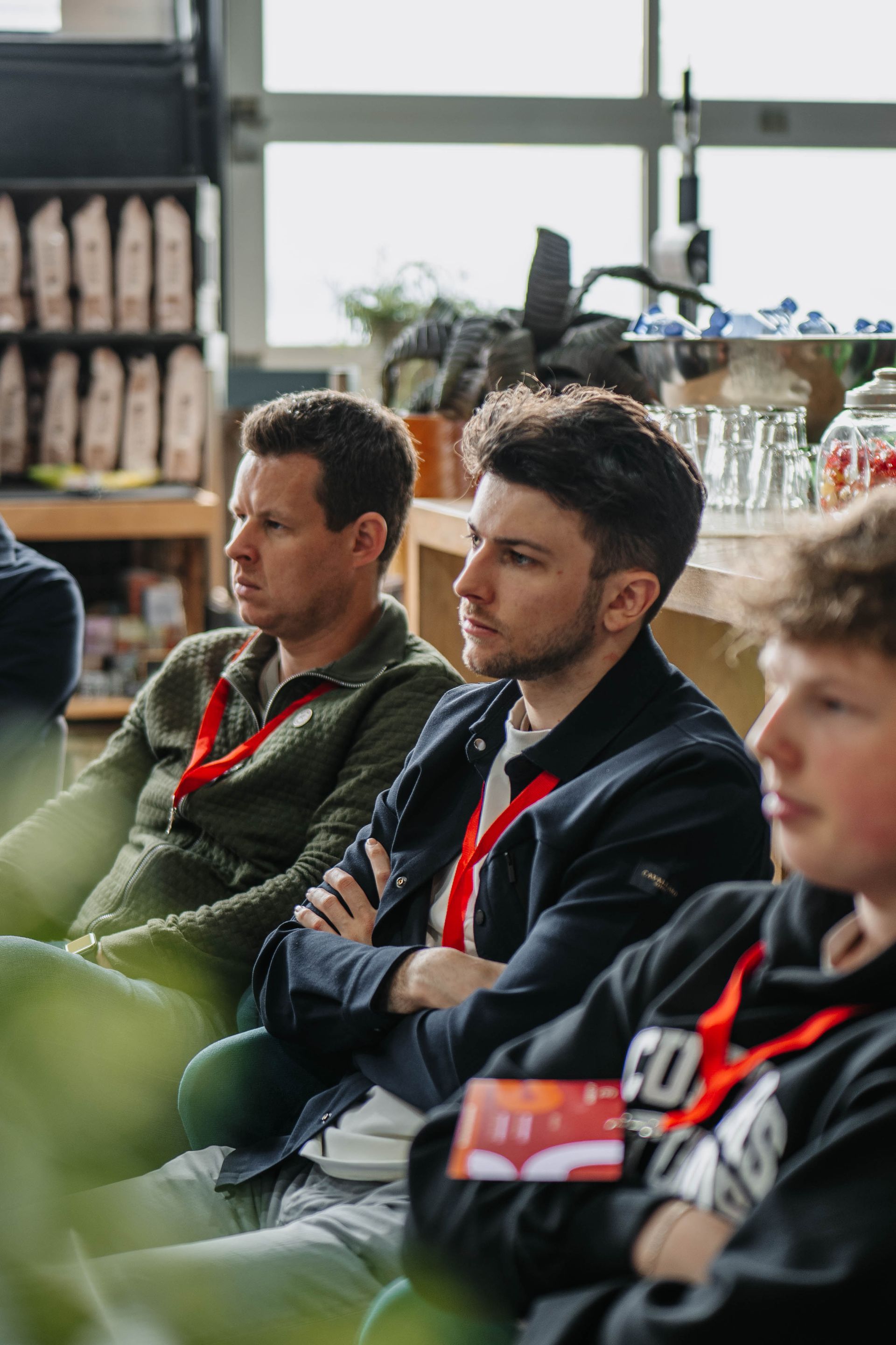 Drie personen, die een conferentiekoord dragen, luisteren aandachtig in een lichte, informele ruimte met planken en glaswerk.