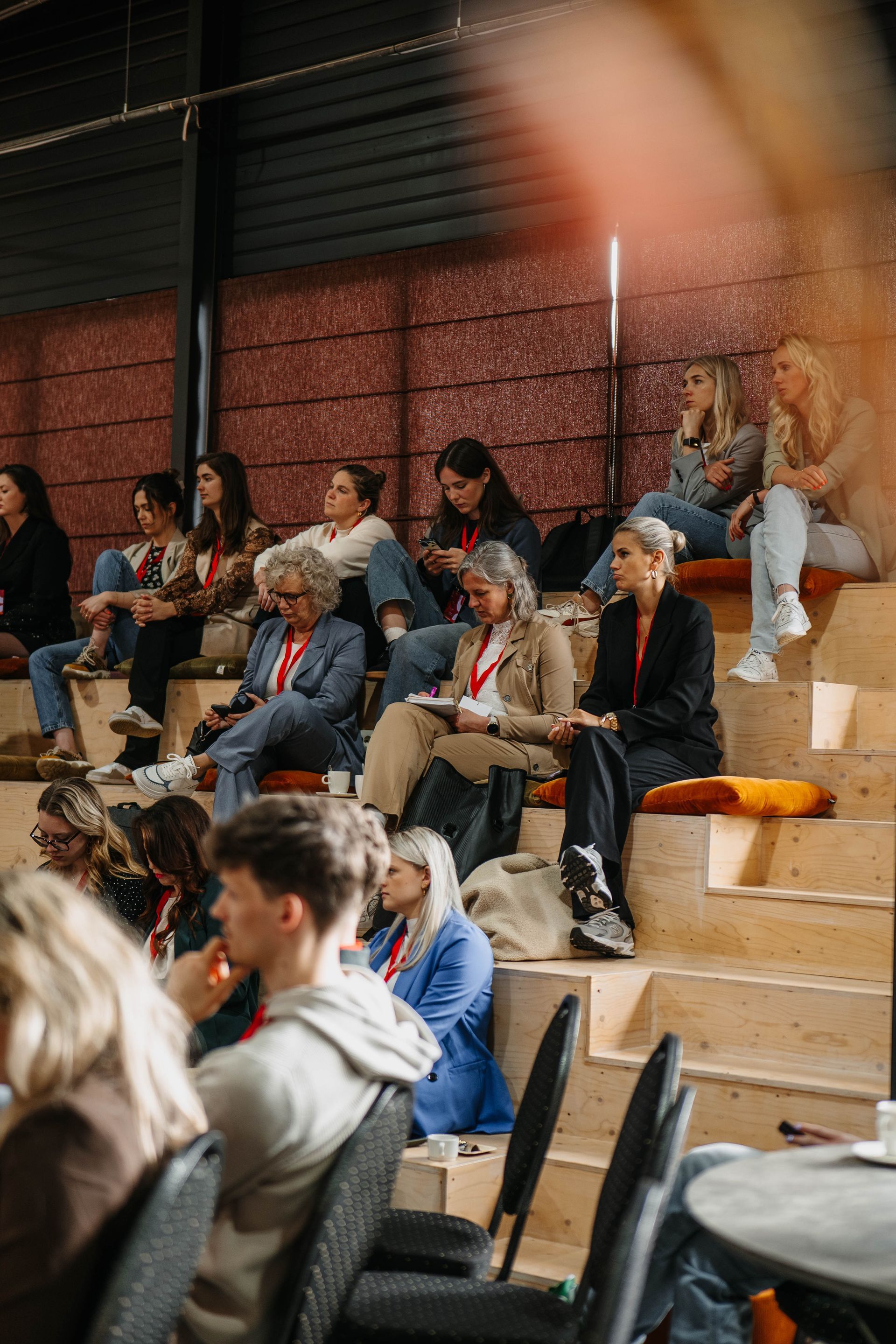 Het publiek zit op een oplopende houten tribune in een zaal met rode, gestructureerde muren en luistert naar een presentatie.