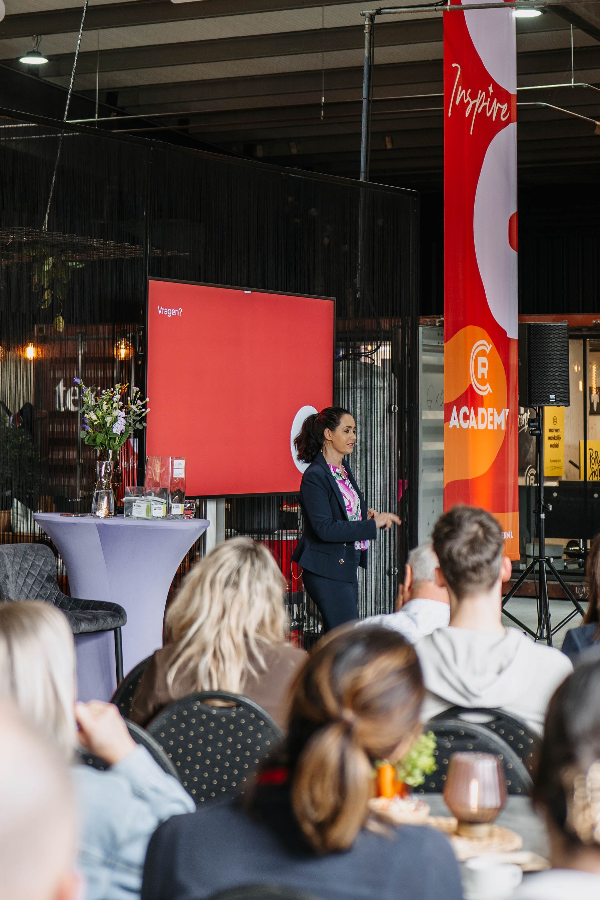 Een spreker in een donkere blazer spreekt een publiek toe in een zaal met grote rode spandoeken met logo.