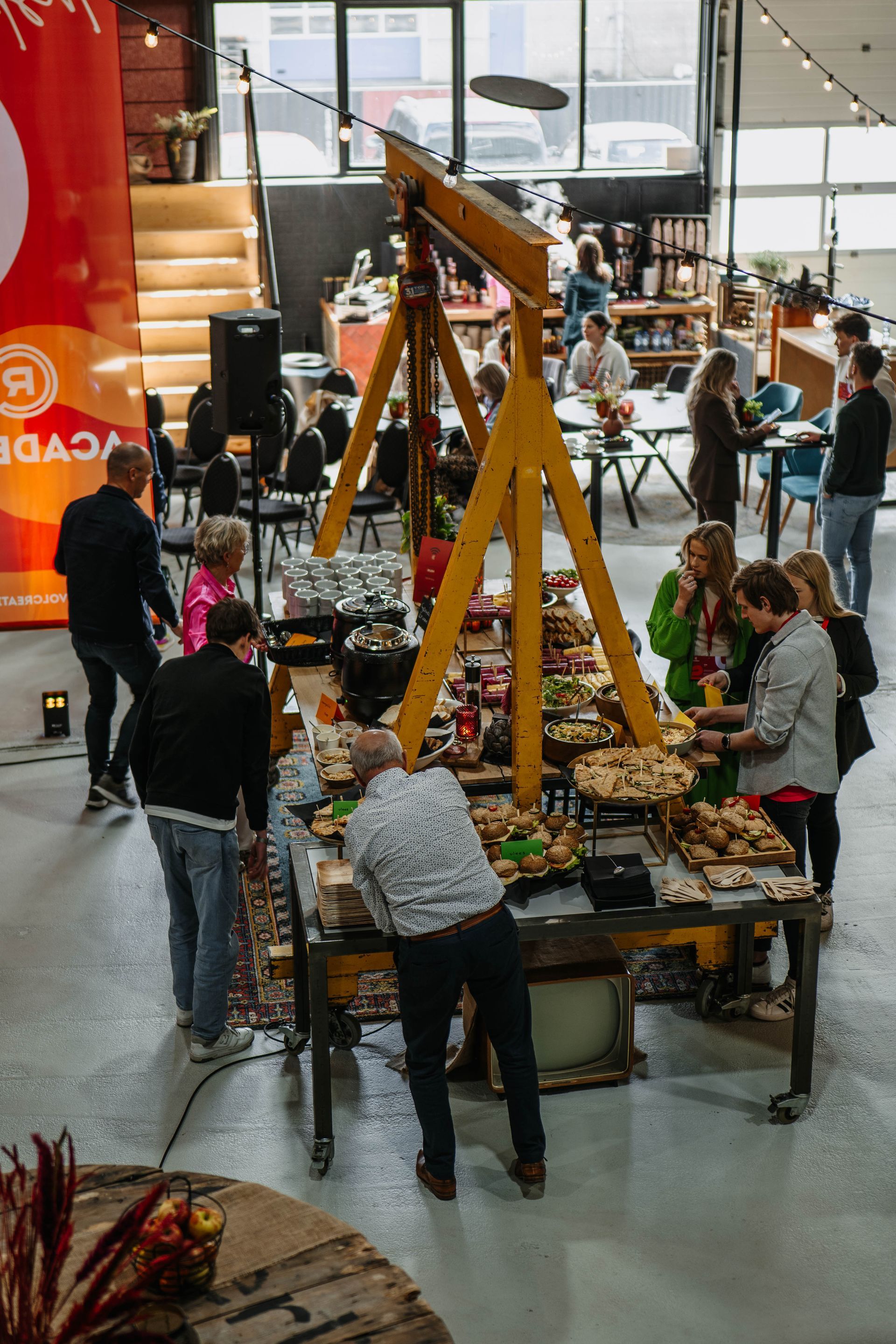 Mensen verzamelen zich rond een buffettafel onder een gele industriële portaalkraan in een felverlichte loods.