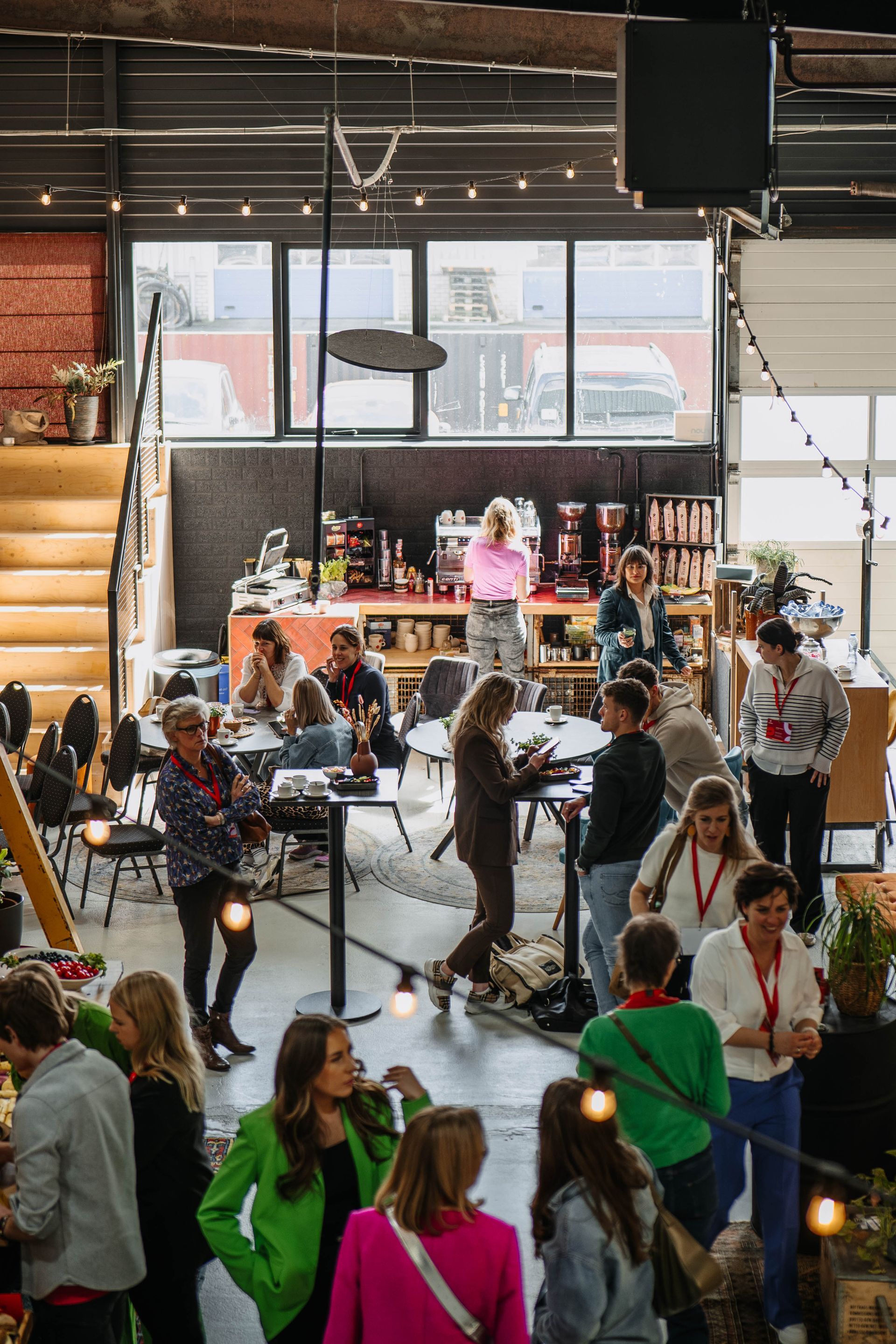 Mensen netwerken en voeren gesprekken in een grote, industriële loods vol tafels, stoelen en sfeervolle verlichting.
