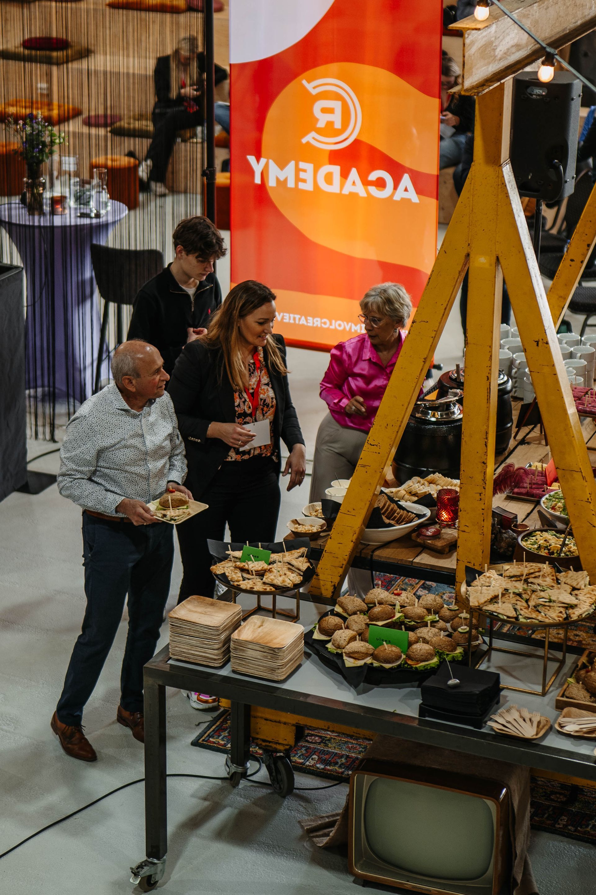 Bezoekers van een evenement staan ​​bij een tafel met hapjes voor een oranje spandoek met het logo van de Academie.