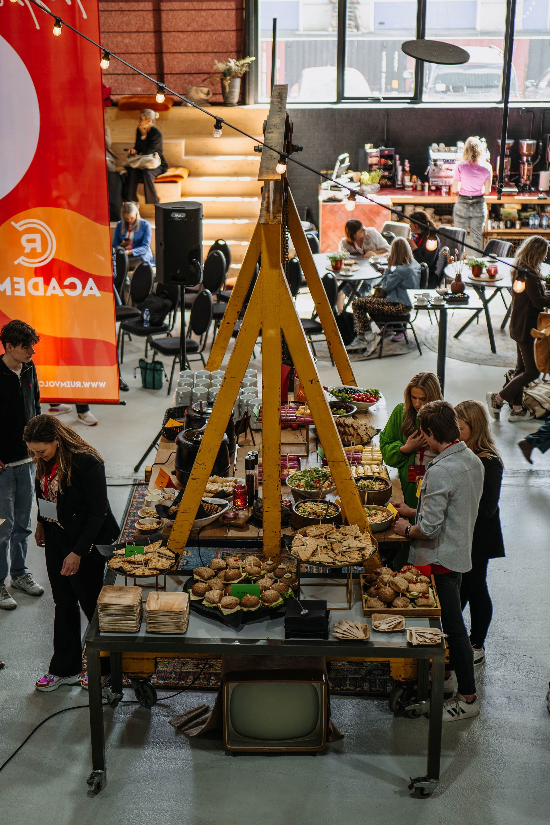 Mensen praten met elkaar rond een grote buffettafel met een gele A-vormige constructie in een lichte, industriële evenementenruimte.