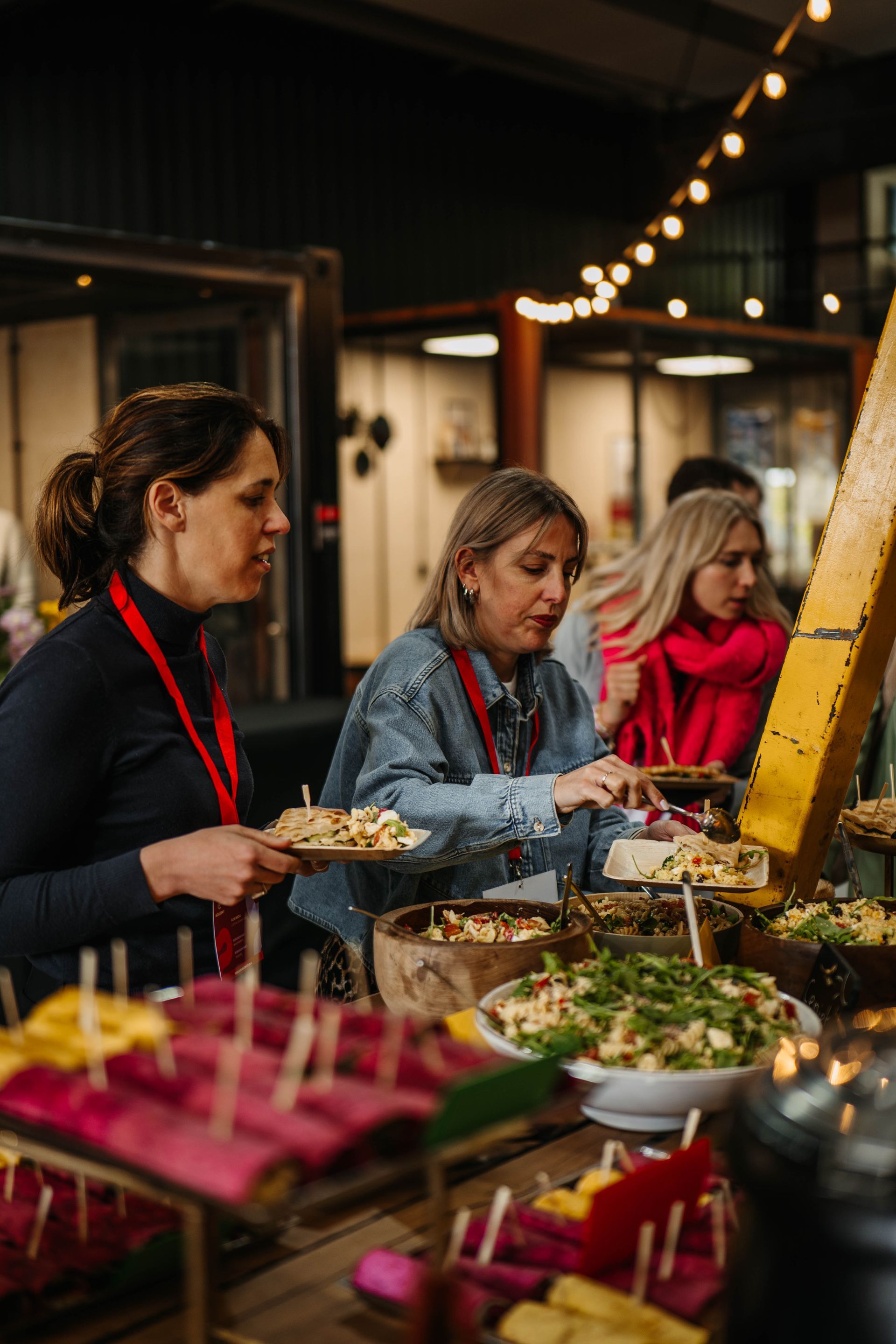 Mensen verzamelen zich rond een buffettafel en kiezen hun gerechten van verschillende schalen en kommen in een sfeervol verlichte, informele evenementenruimte.