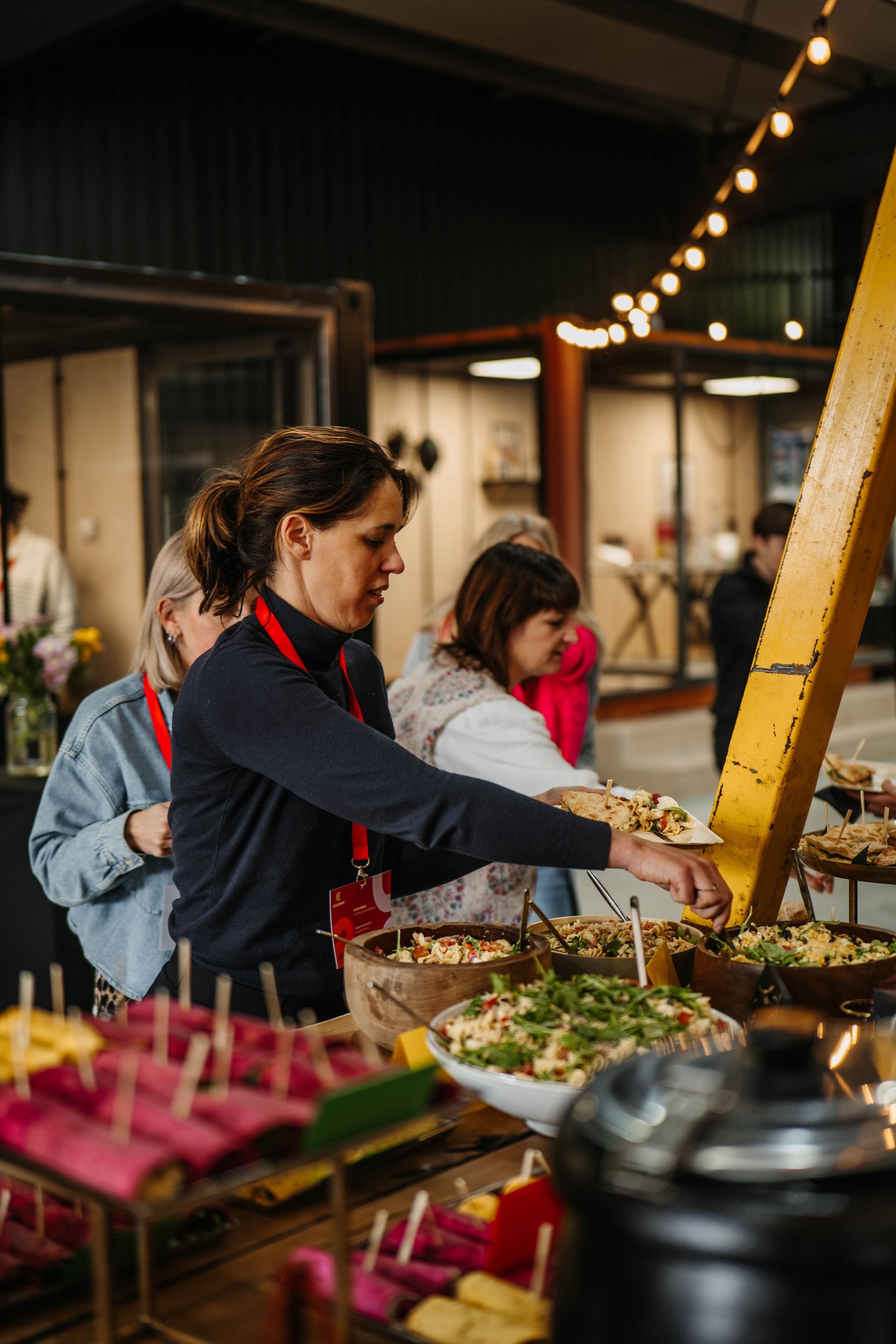 Een groep mensen staat in de rij bij een buffet en schept zelf eten op uit verschillende schalen tijdens een evenement binnenshuis.