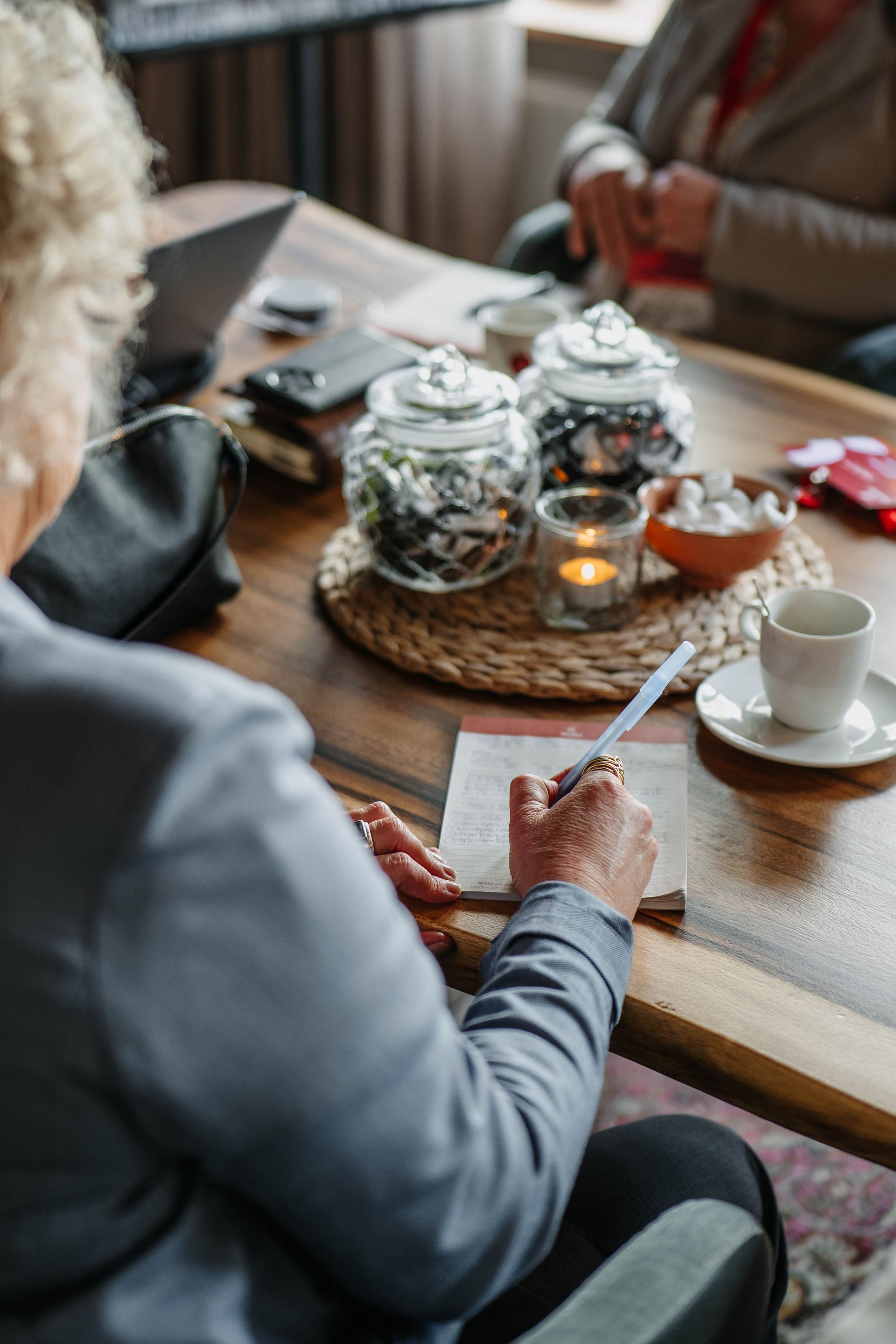 Een persoon in een grijze blazer schrijft op papier aan een houten tafel met glazen potten, een brandende kaars en een koffiekopje.