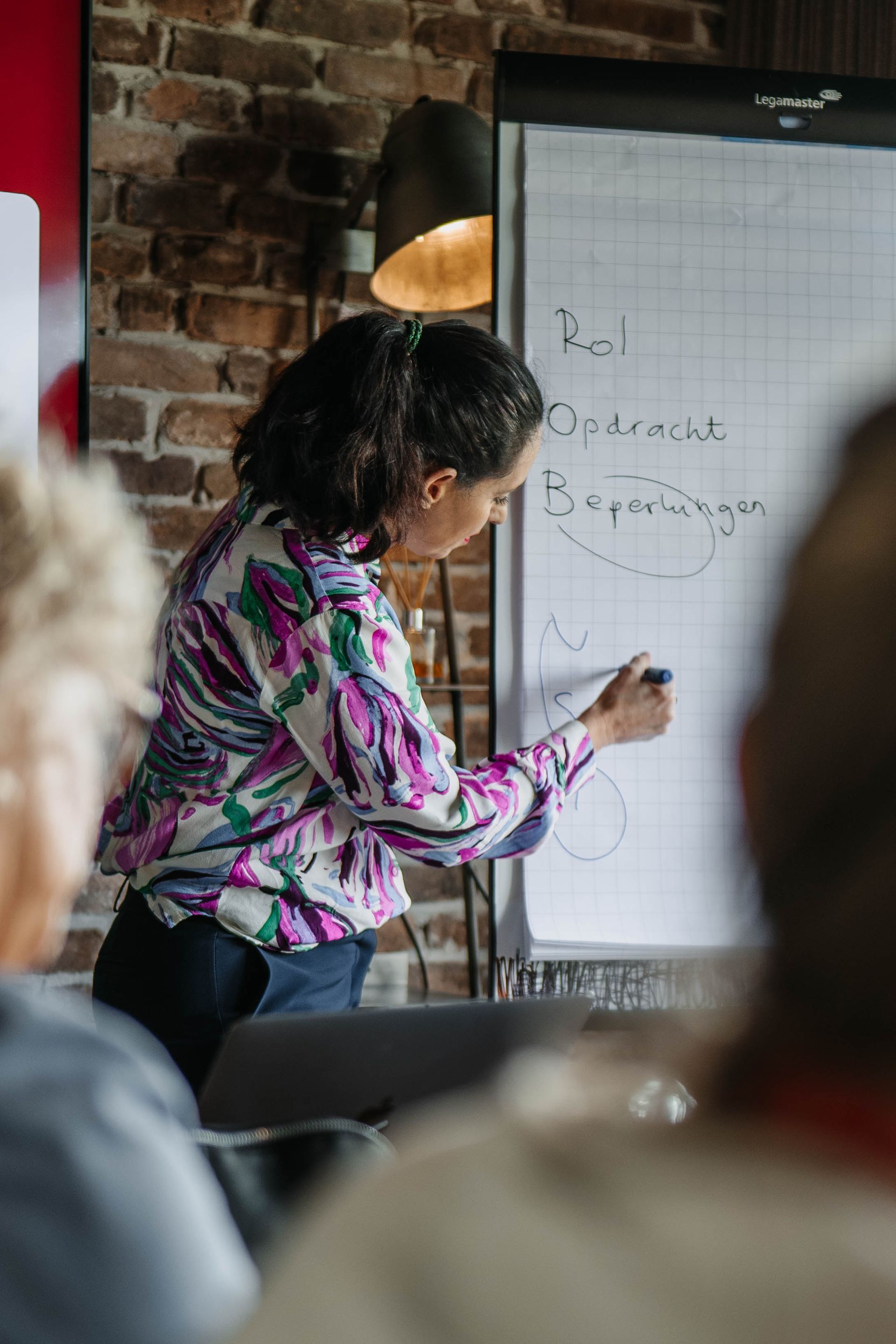 Een persoon in een overhemd met een patroon schrijft op een flipchart in een ruimte met een bakstenen muur, gezien vanaf achter een publiek.