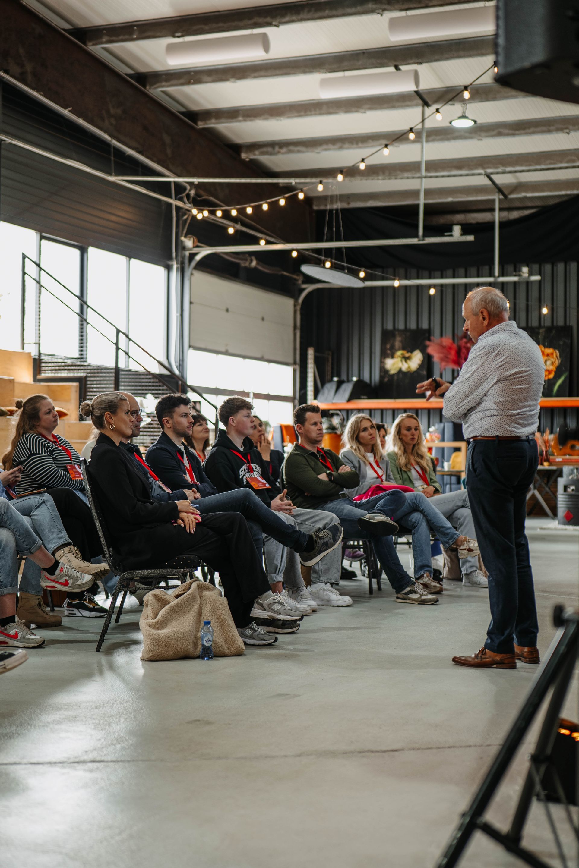 Een spreker staat voor een publiek in een industriële ruimte en spreekt een groep toe die op stoelen zit.