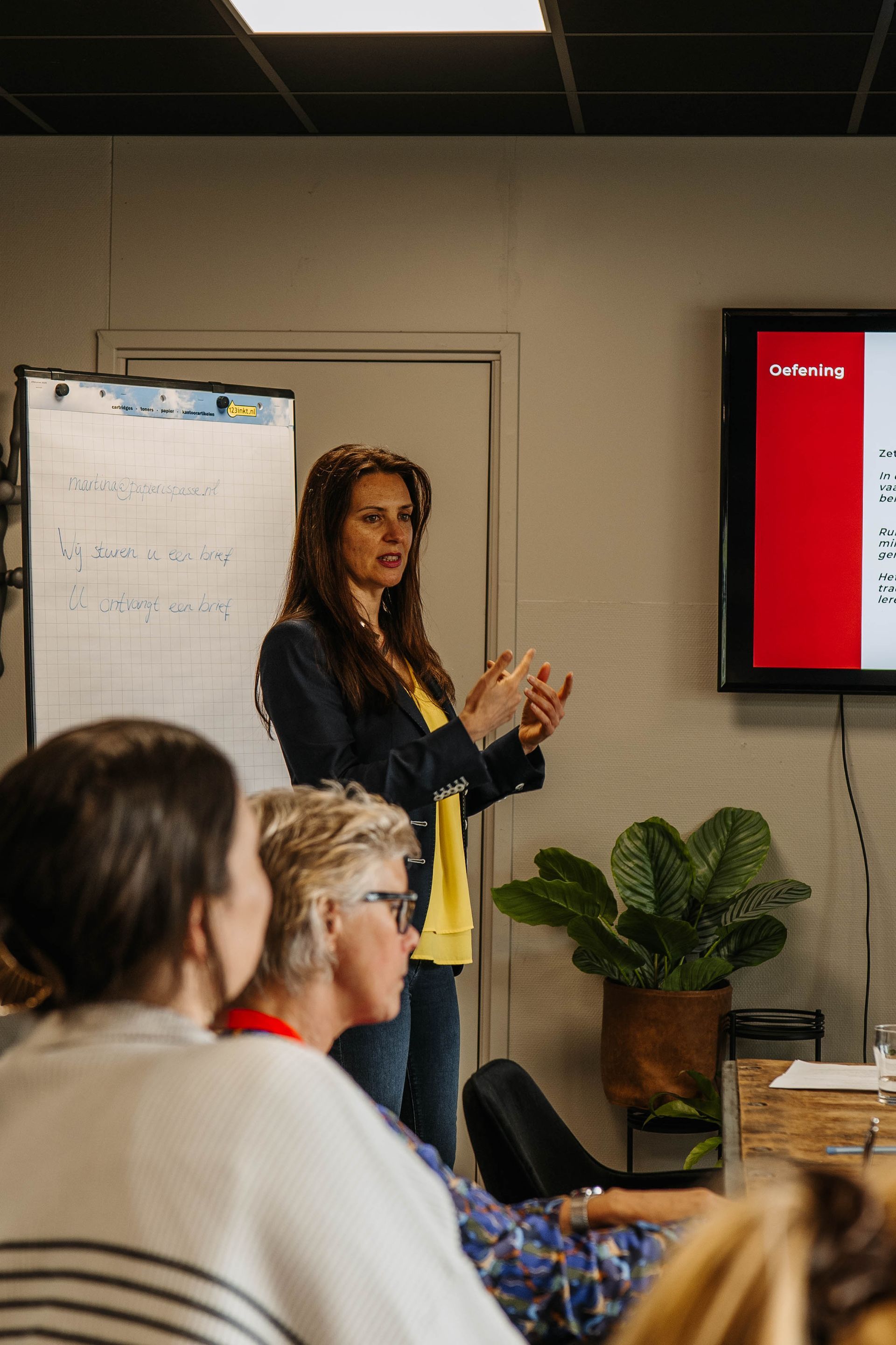 Een vrouw in een gele top en een donkere jas spreekt een publiek toe in een kantooromgeving met een whiteboard en een digitaal scherm.