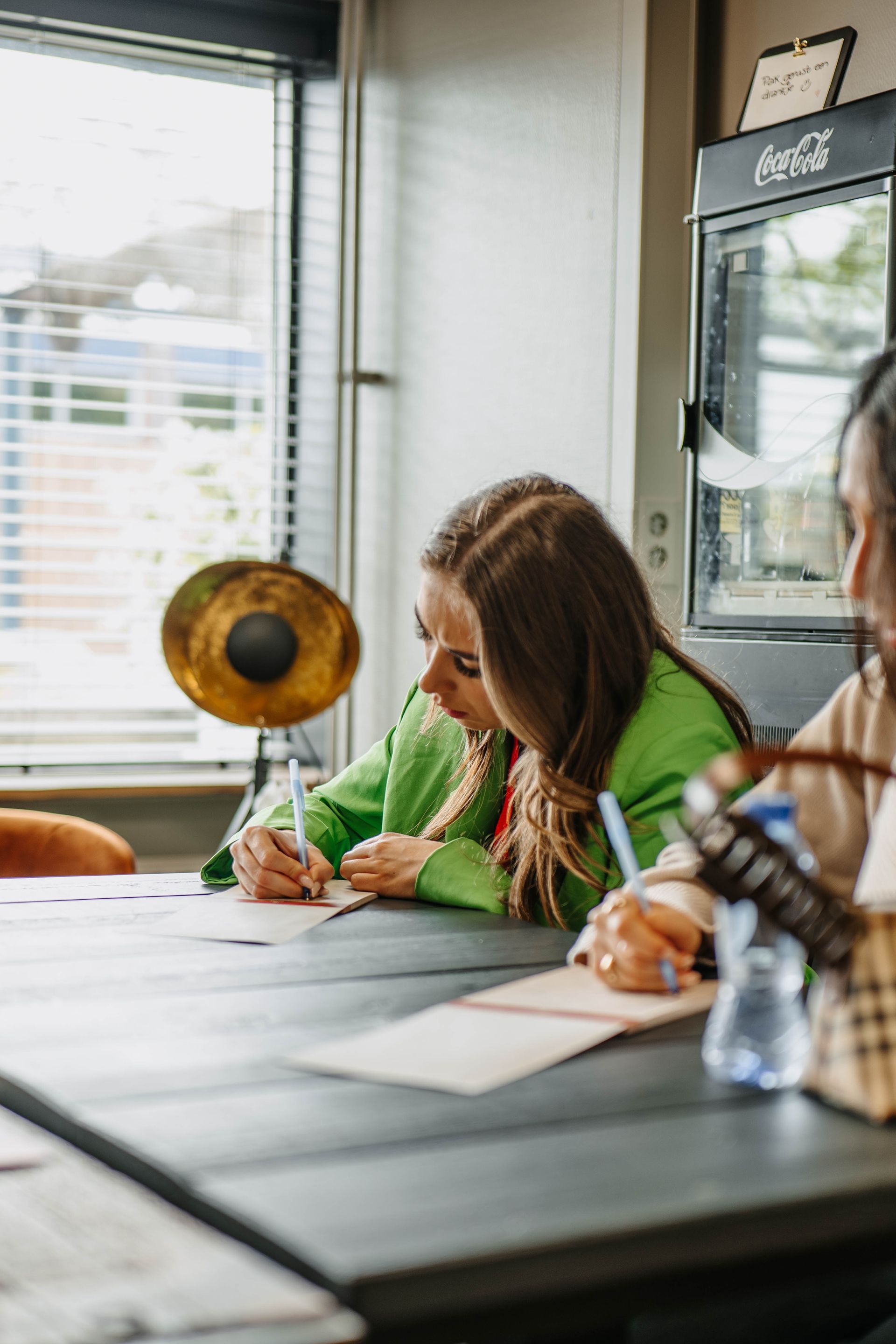 Twee mensen zitten aan een tafel te schrijven in notitieboekjes in een goed verlichte kamer met een raam en een Coca-Cola-koelkast in de buurt.
