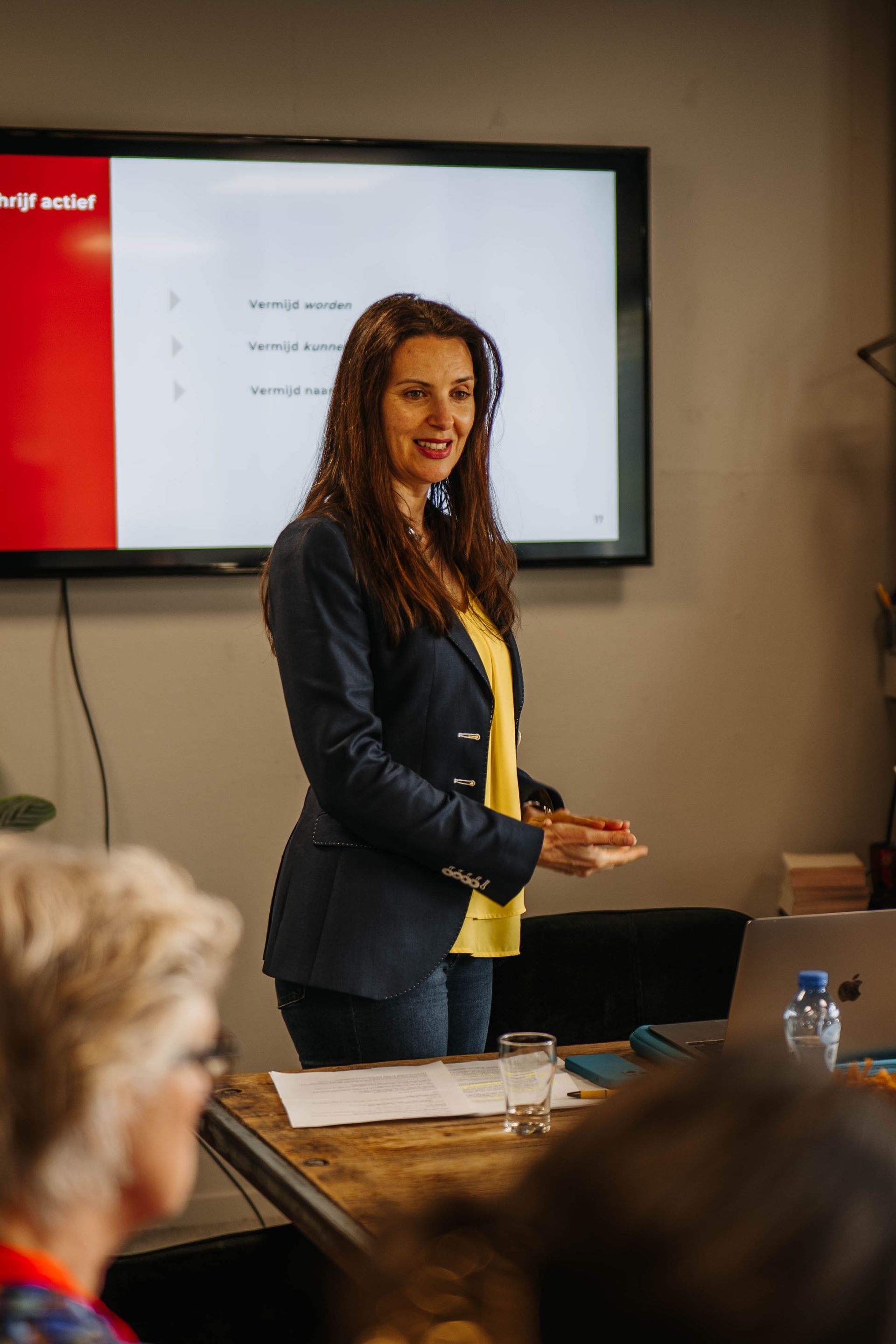 Een presentator in een donkerblauwe blazer en een gele top staat voor een presentatiescherm in een kamer met een houten tafel.