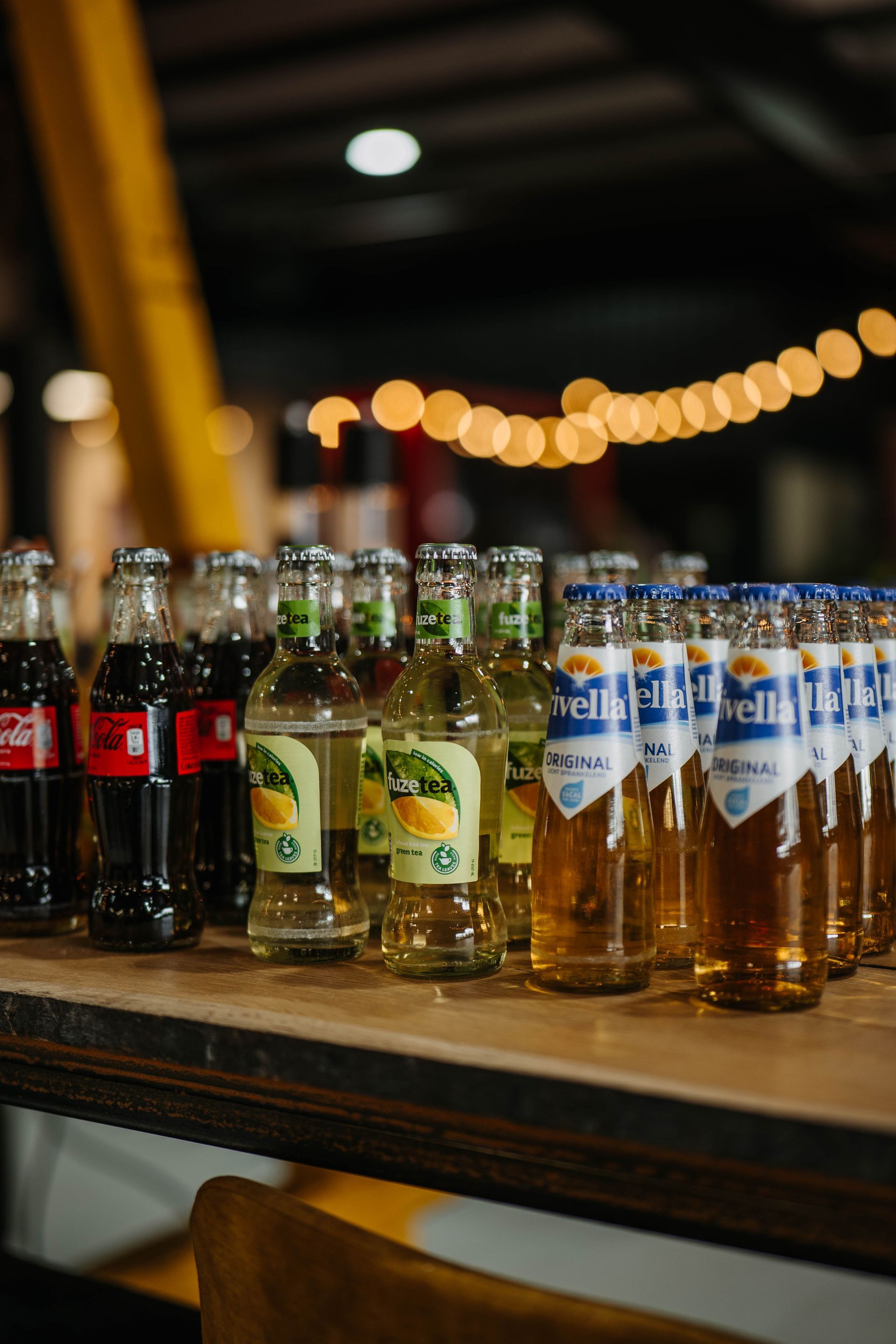 Een rij diverse frisdrank- en bierflessen op een houten toonbank, met wazige lichtslingers op de achtergrond.