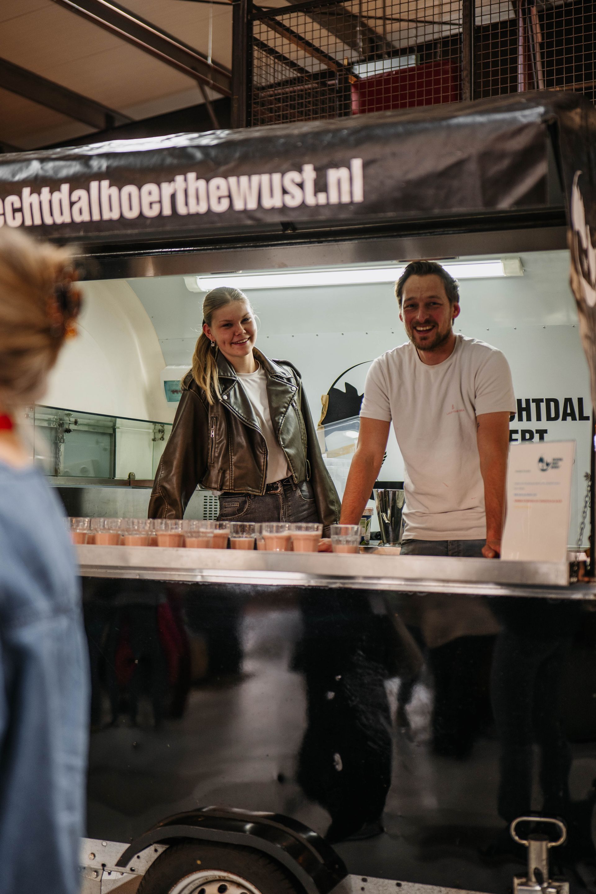 Twee lachende mensen serveren voedselmonsters vanuit een kraam met het opschrift 
