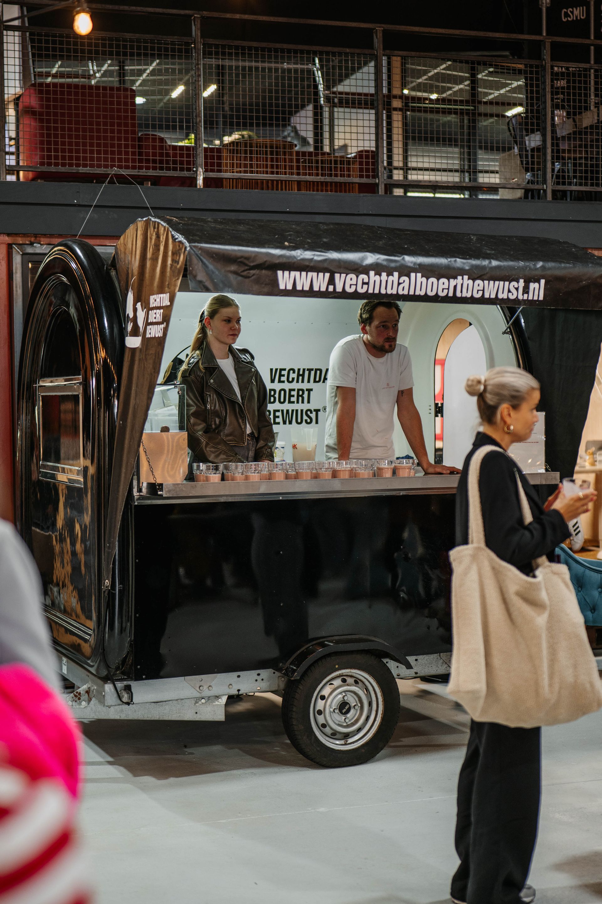 Twee mensen werken achter een zwarte cateringtrailer bij een evenement binnenshuis, met een klant op de voorgrond.