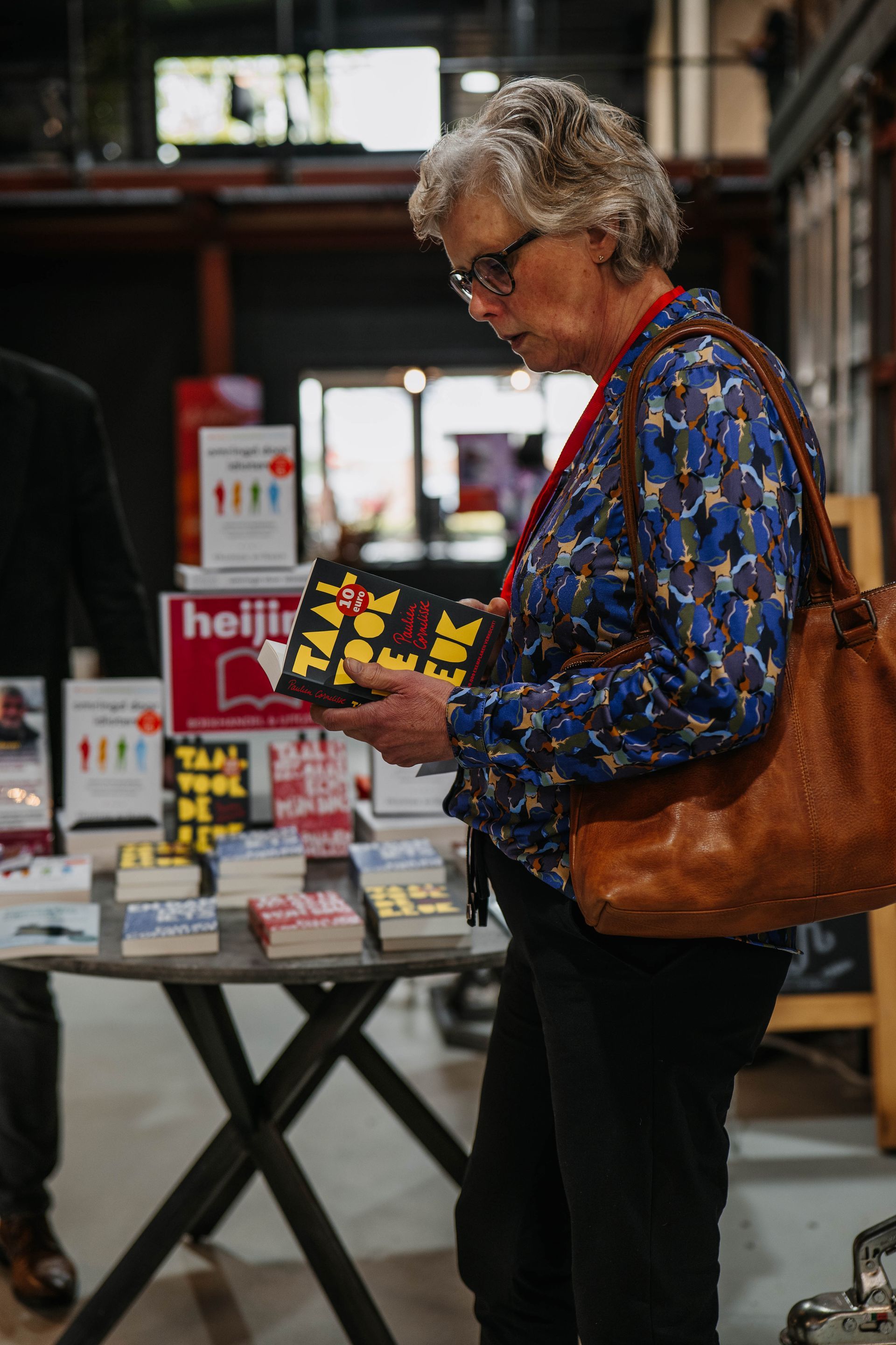 Een persoon in een blauw overhemd met patroon bekijkt een zwart boek terwijl hij aan een tafel staat waarop verschillende boeken uitgestald staan.