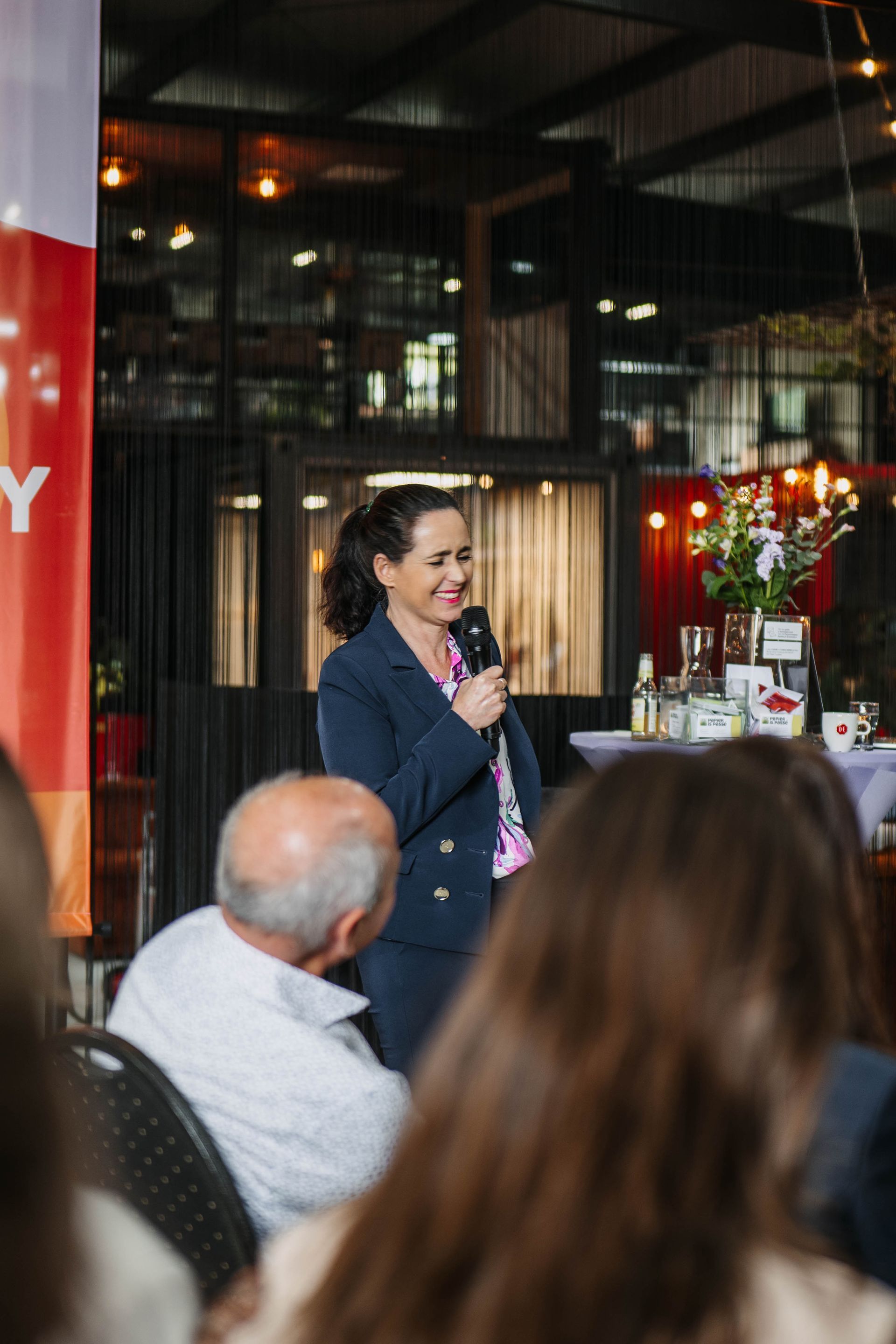 Een vrouw in een donkerblauw pak spreekt in een microfoon tijdens een evenement binnenshuis, terwijl ze glimlachend naar het publiek kijkt.