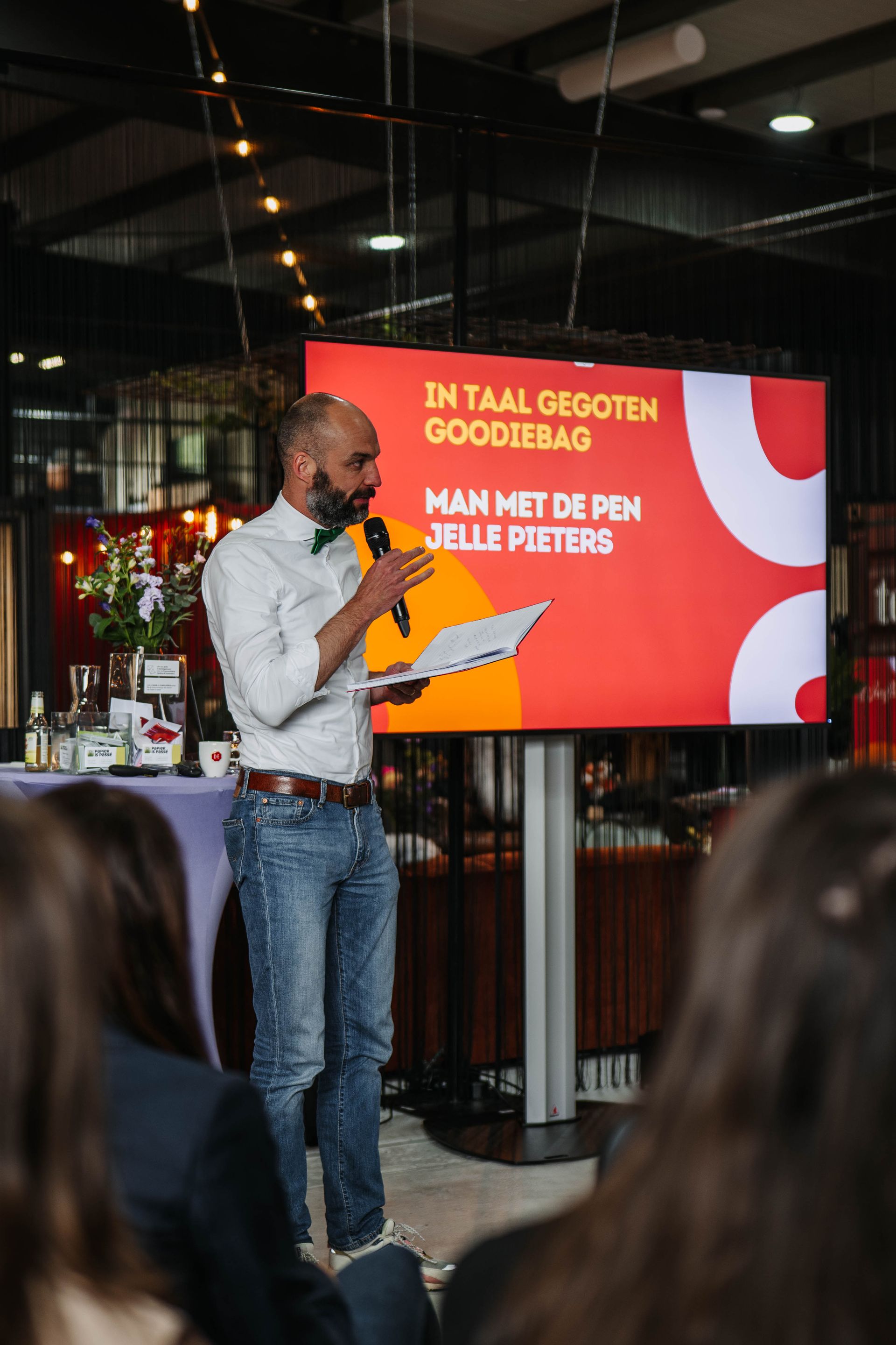 Een persoon in een wit shirt en spijkerbroek spreekt vanaf een podium voor een rood scherm met de titel 