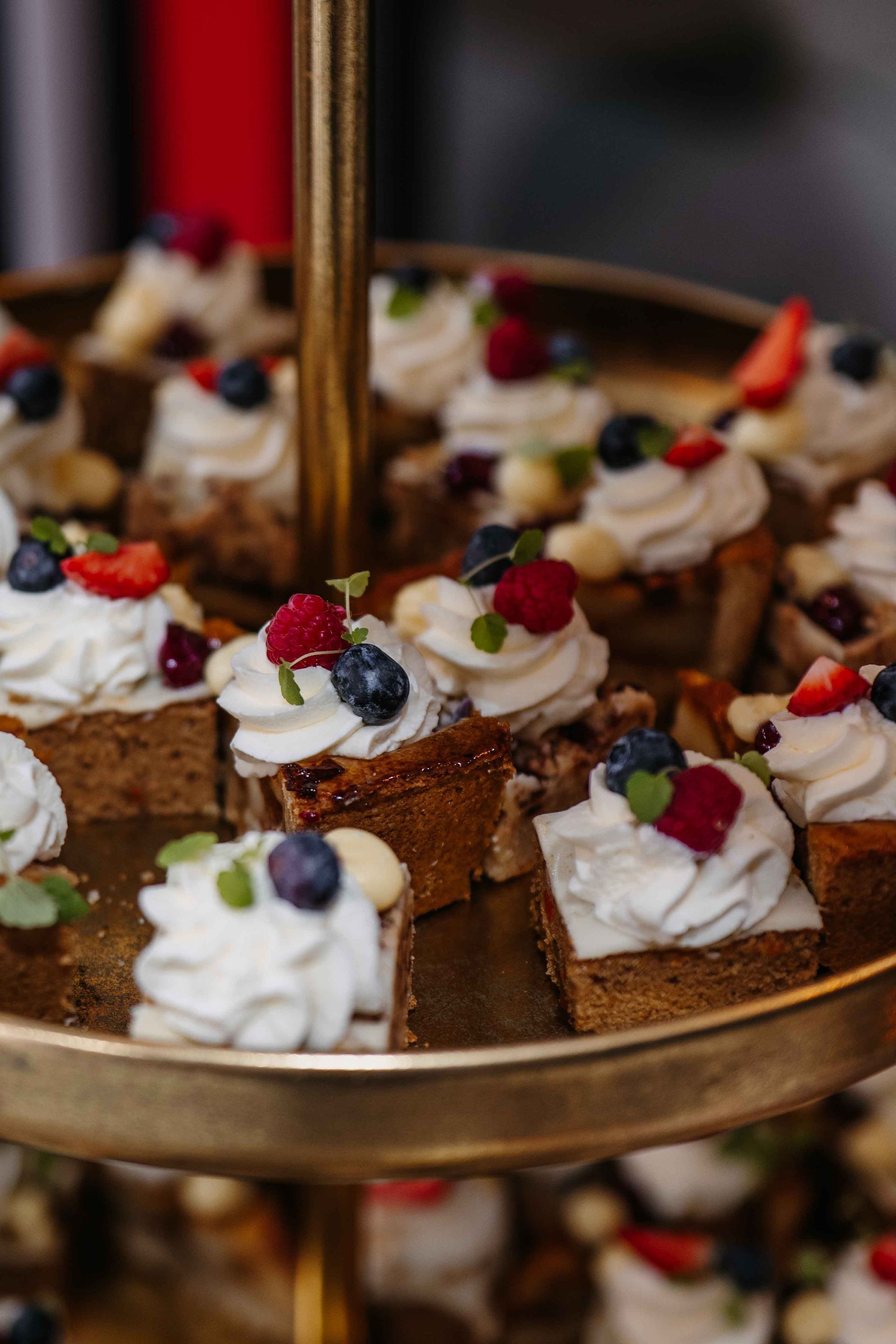 Een gouden, tweelaags dessertetagère gevuld met individuele vierkante gebakjes, gegarneerd met witte slagroom, bosbessen en frambozen.