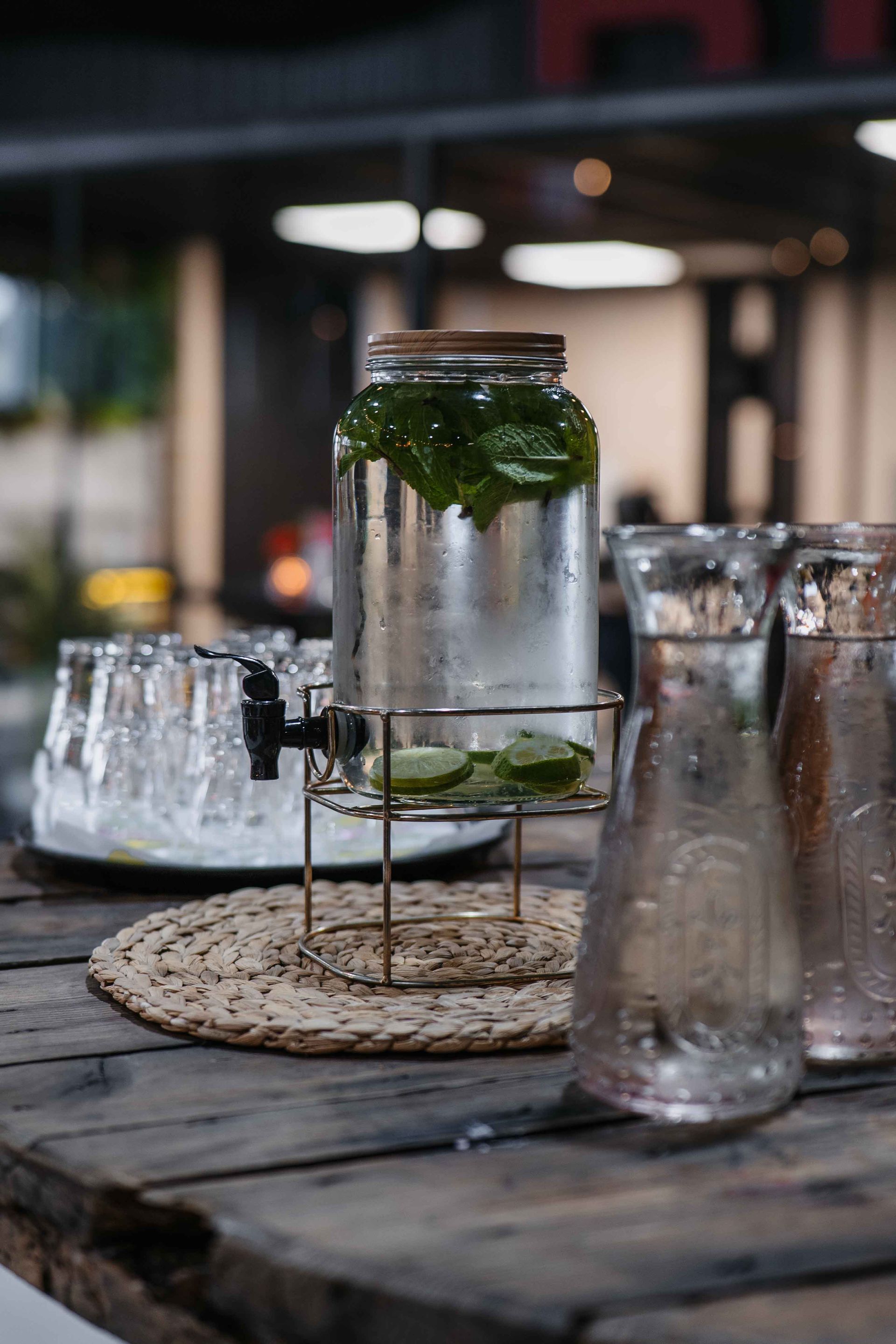 Een drankdispenser gevuld met munt- en limoenwater staat op een geweven mat naast een dienblad met glazen op een houten tafel.