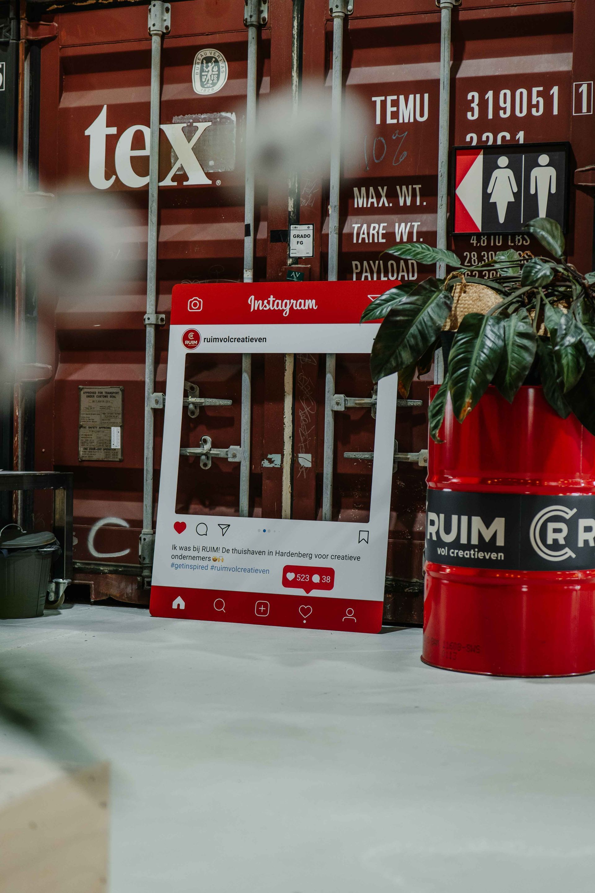 Een rood fotolijstje in Instagram-stijl staat tegen een zeecontainer naast een rood vat met een groene plant.
