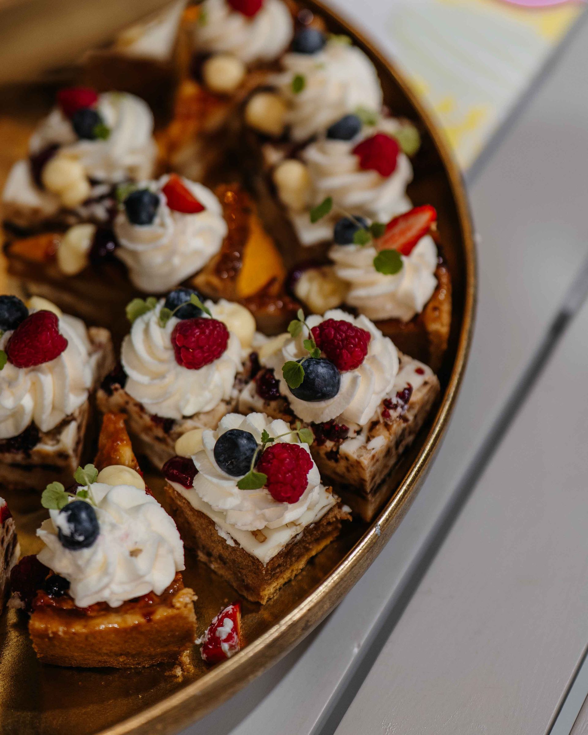 Een gouden schaal met diverse kleine, vierkante desserthapjes, gegarneerd met witte room, bosbessen, frambozen en munt.