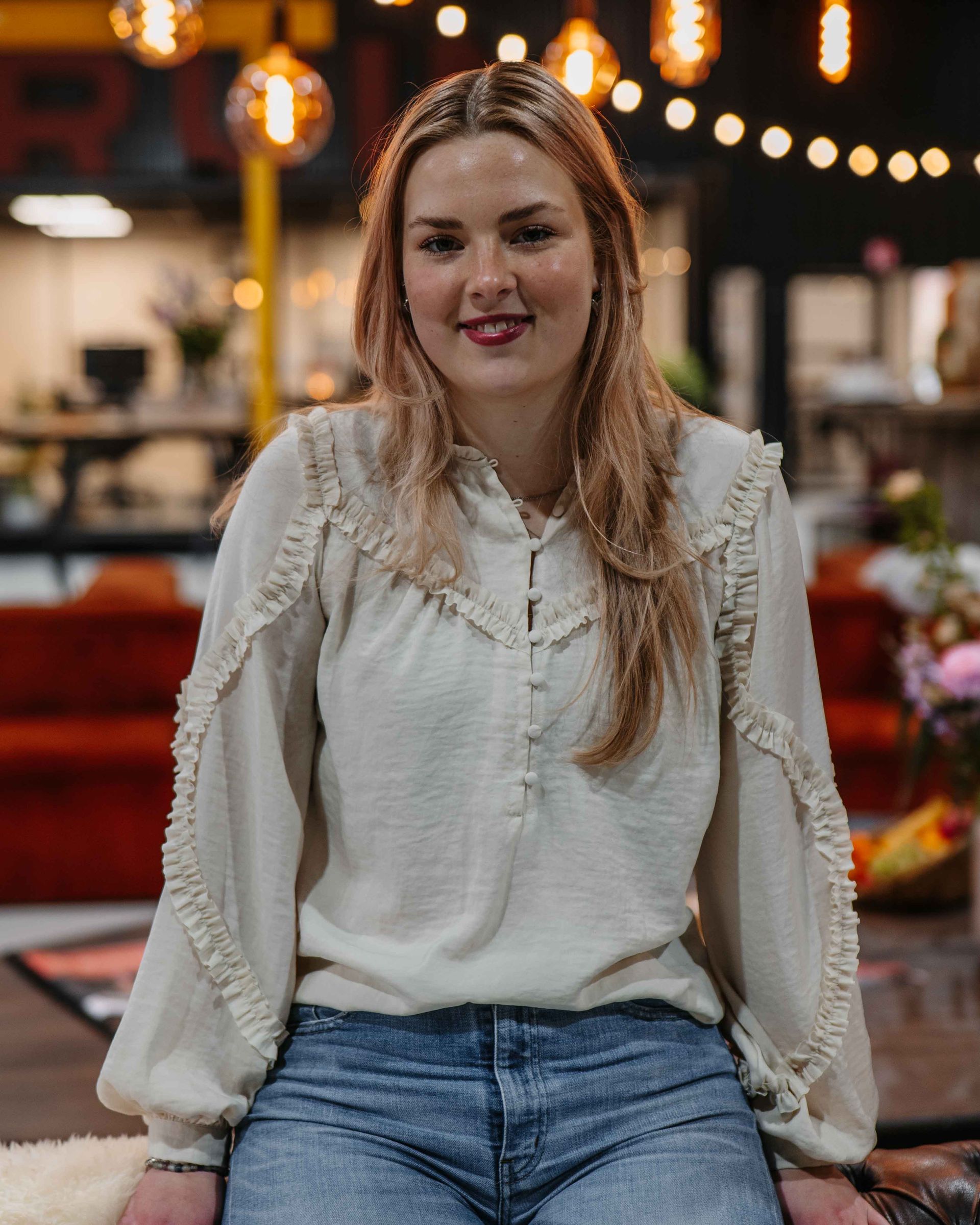 Een glimlachend persoon draagt ​​een crèmekleurige, gegolfde blouse met lange mouwen en een blauwe spijkerbroek in een sfeervol verlichte, overdekte evenementenruimte.